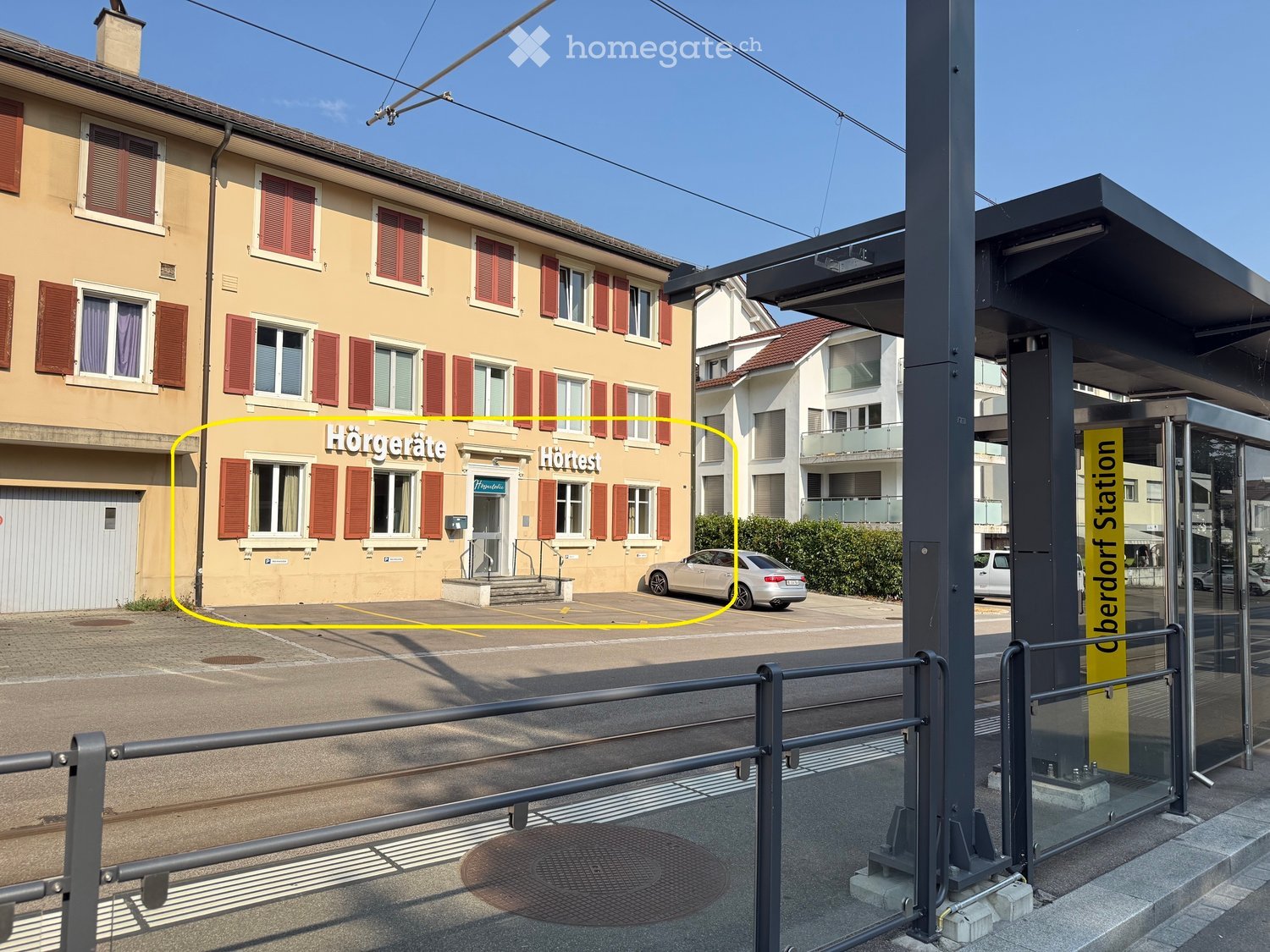 A multi-story building with a beige facade, red shutters, and a sign reading "Hörgeräte" and "Hörest". There is a parking area in front with a silver car and a bus stop shelter to the right.