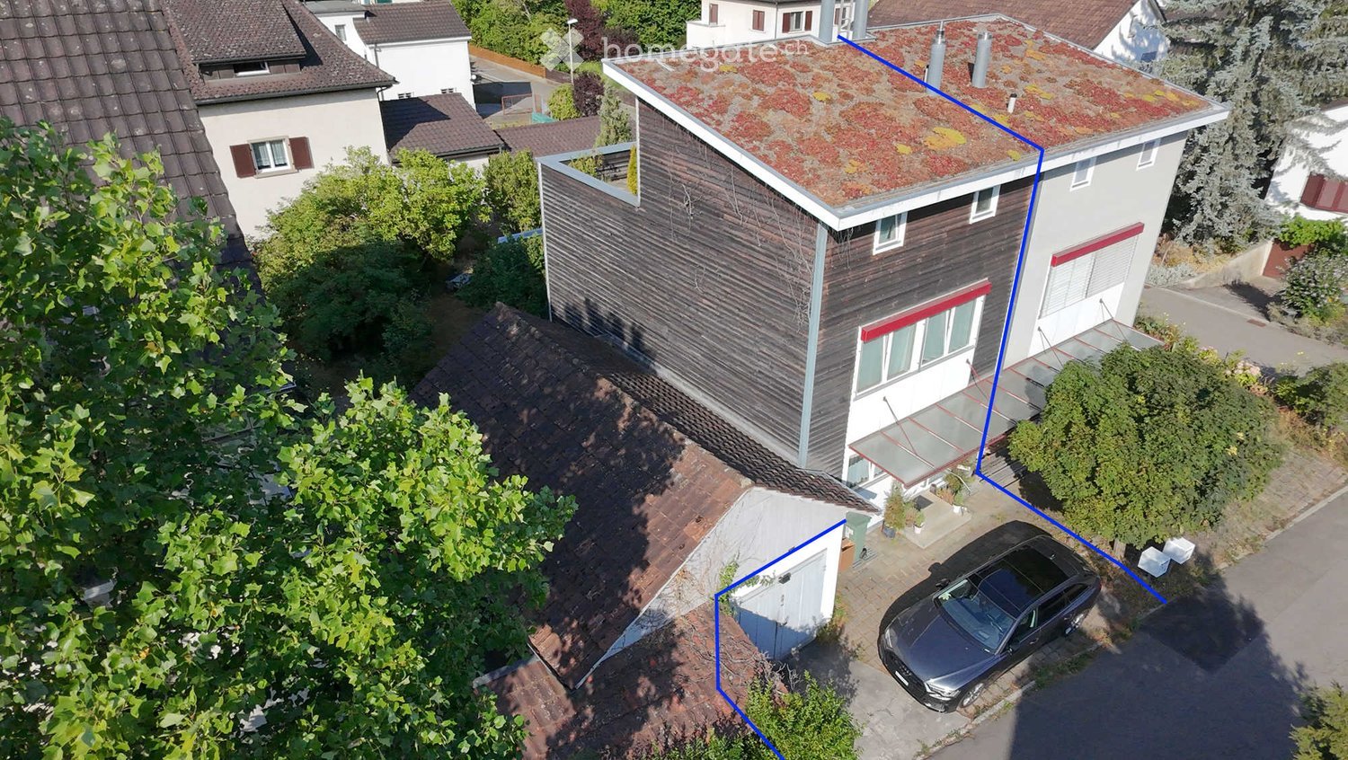 Aerial view of a modern two-story house with a flat roof, surrounded by greenery and neighboring houses. A car is parked in the driveway, and there is a small garage visible.