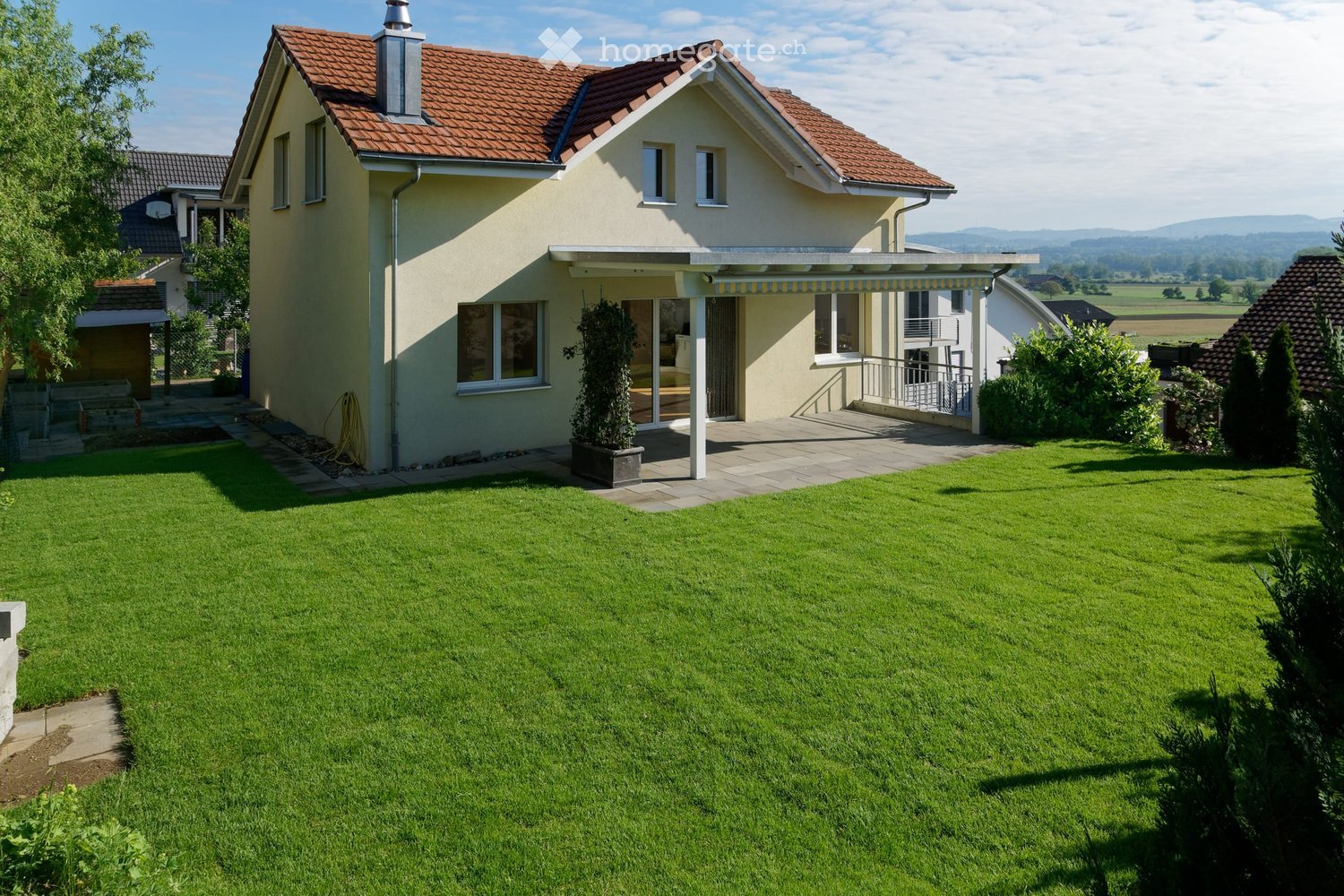 Two-story house with a red-tiled roof, beige exterior, and a well-maintained lawn in the front yard. Features include a covered patio area with a railing and a small garden with a potted plant.