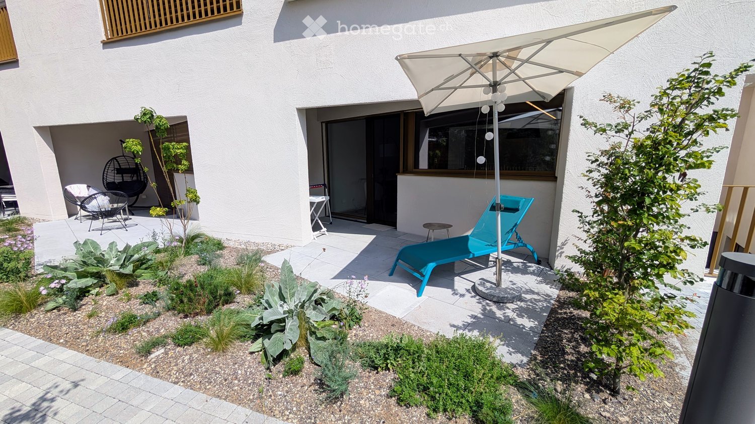 Well-maintained garden area with a patio, lounge chair, umbrella, small seating area, and various plants and shrubs.