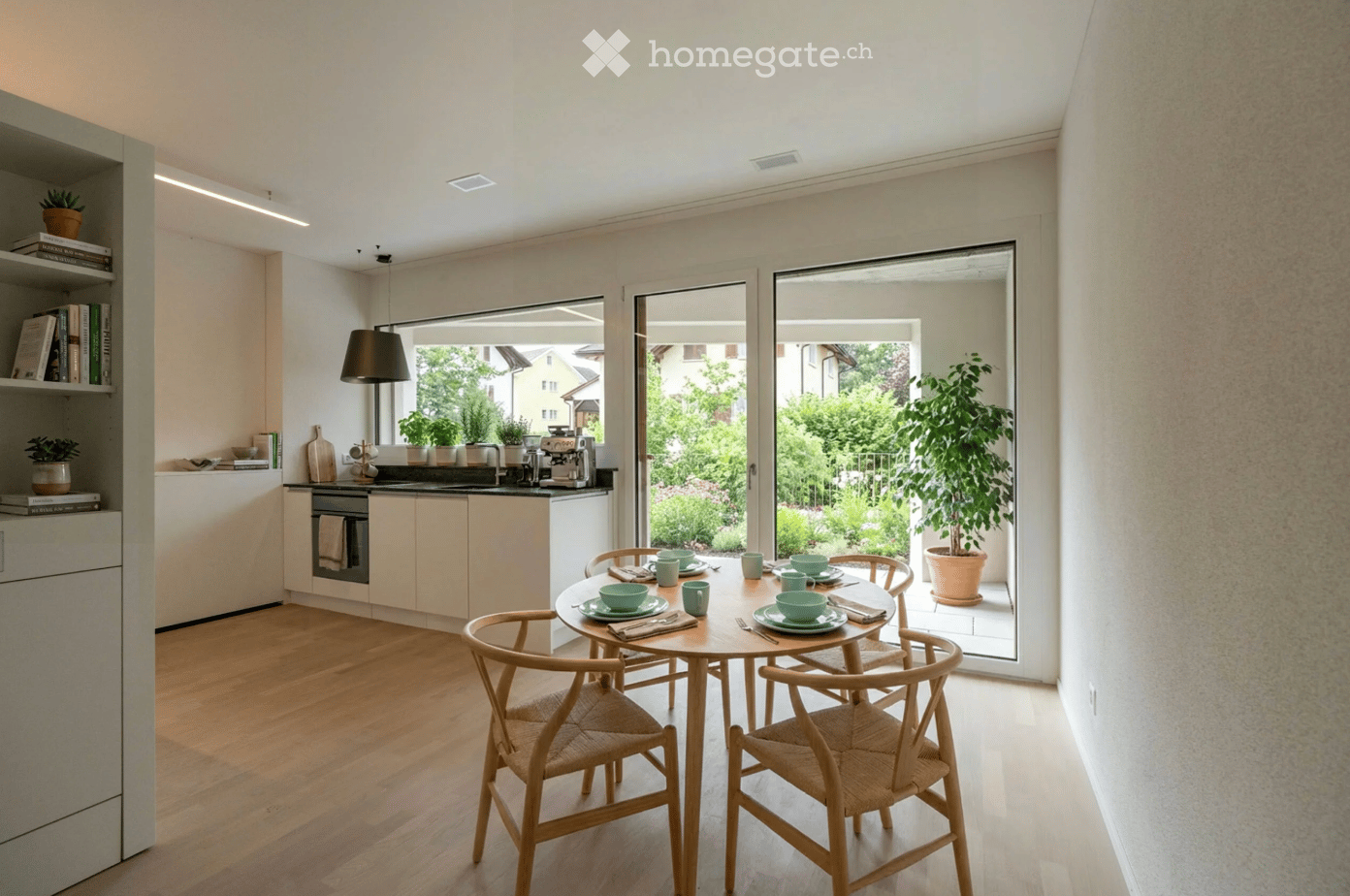 Modern kitchen with light-colored cabinets, a round dining table with four chairs, and a view of a garden through large windows.
