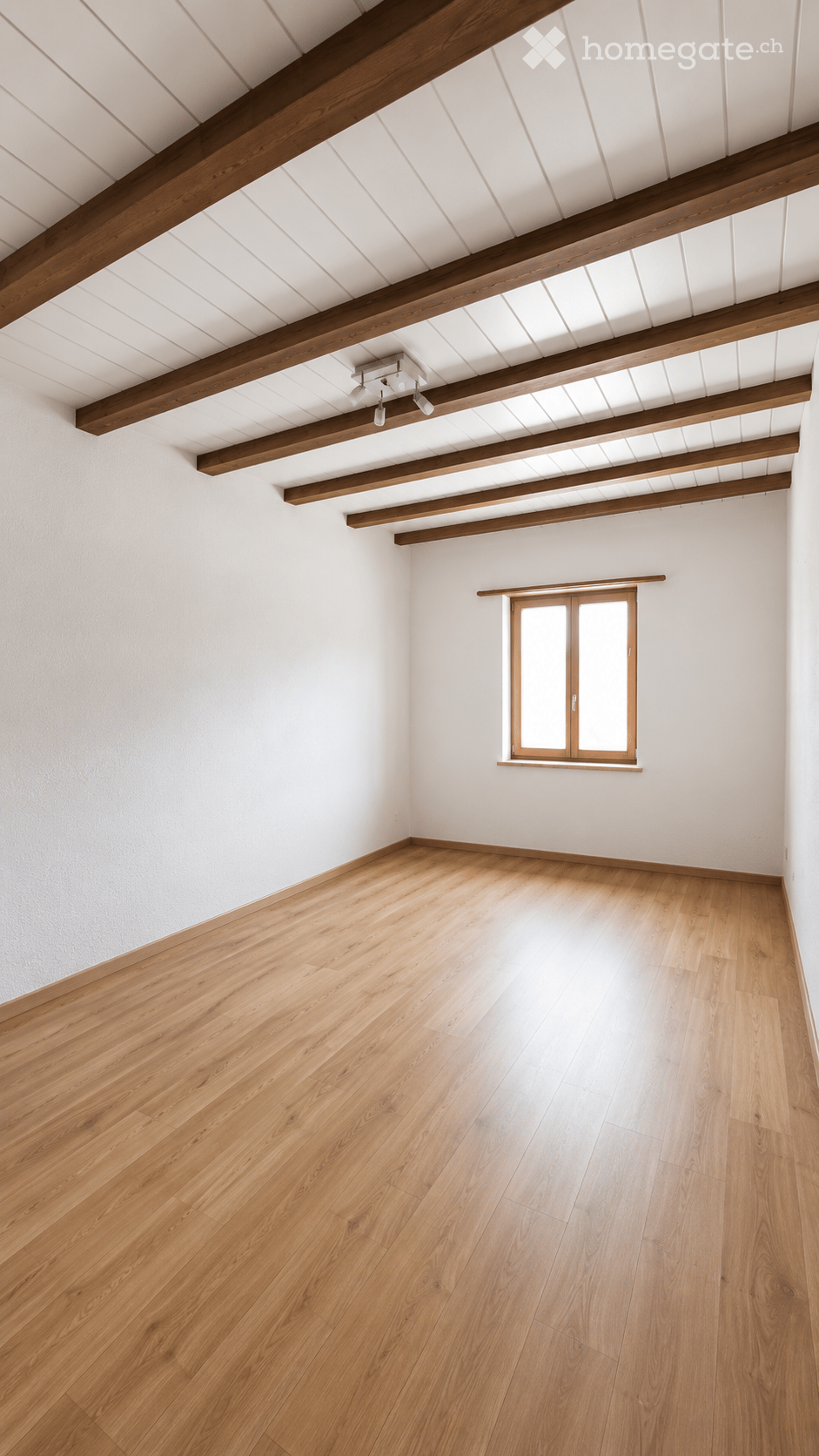 Empty room with wooden flooring, white walls, exposed wooden beams on the ceiling, and a single window with wooden frame.