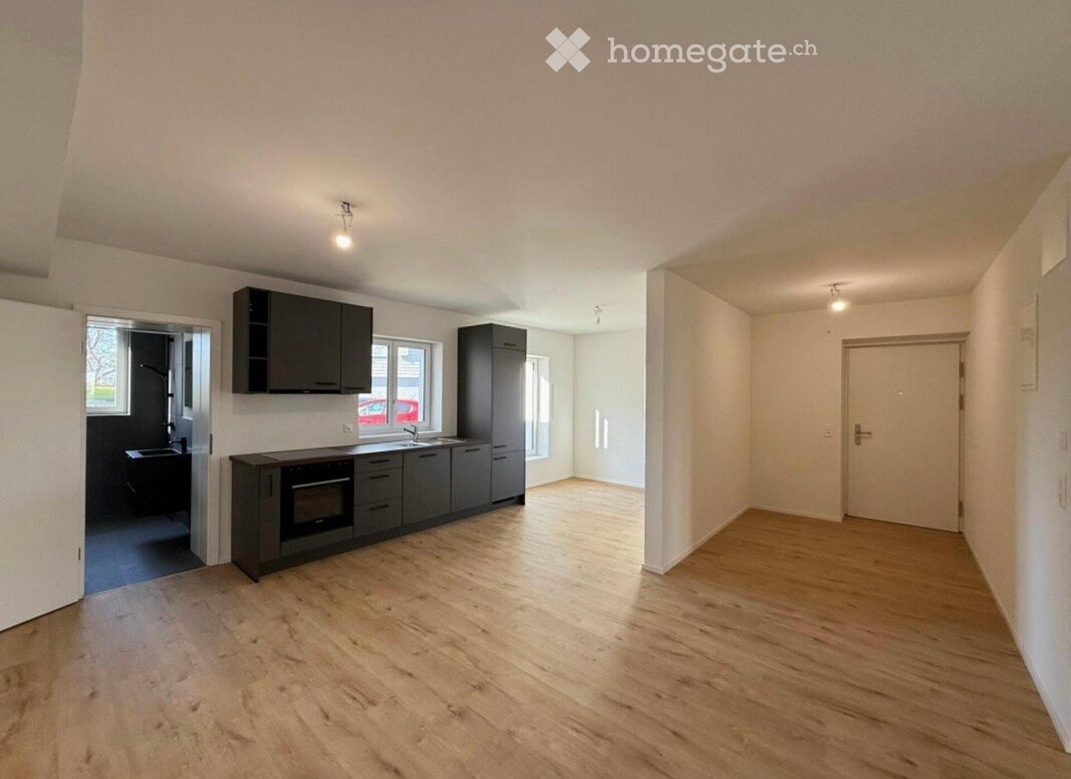 Empty room with light wooden flooring, white walls, and a doorway leading to a kitchen area with dark cabinets and appliances.