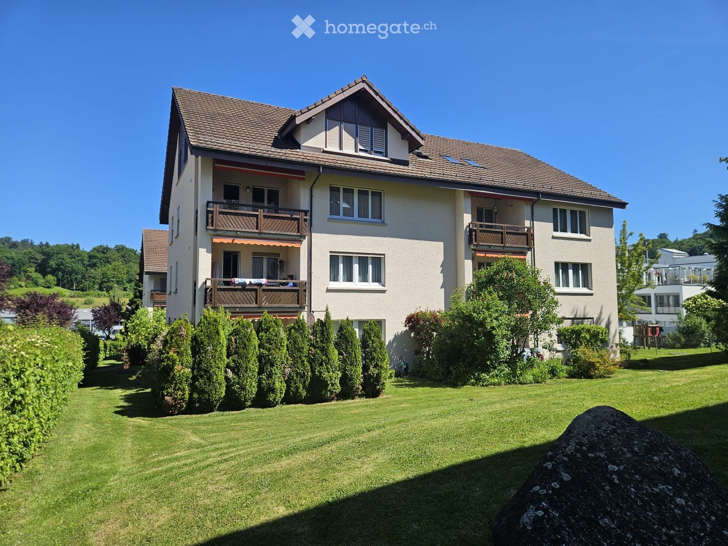 Two-story house with beige walls, brown tiled roof, multiple windows, balconies, and surrounding greenery.