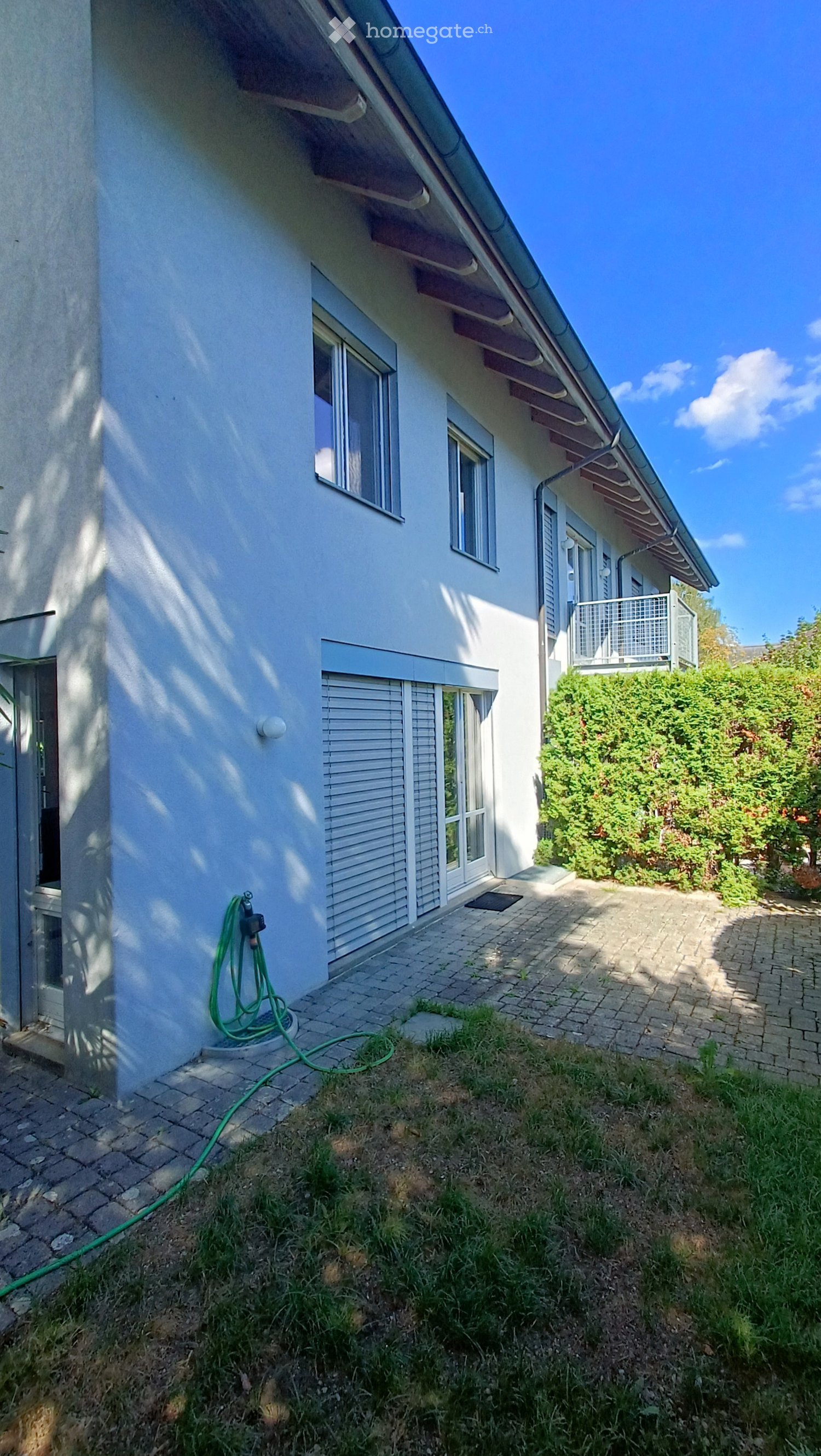 Two-story house with white exterior, multiple windows, and a small balcony on the upper level. The house has a paved pathway leading to the entrance and a garden area with greenery.