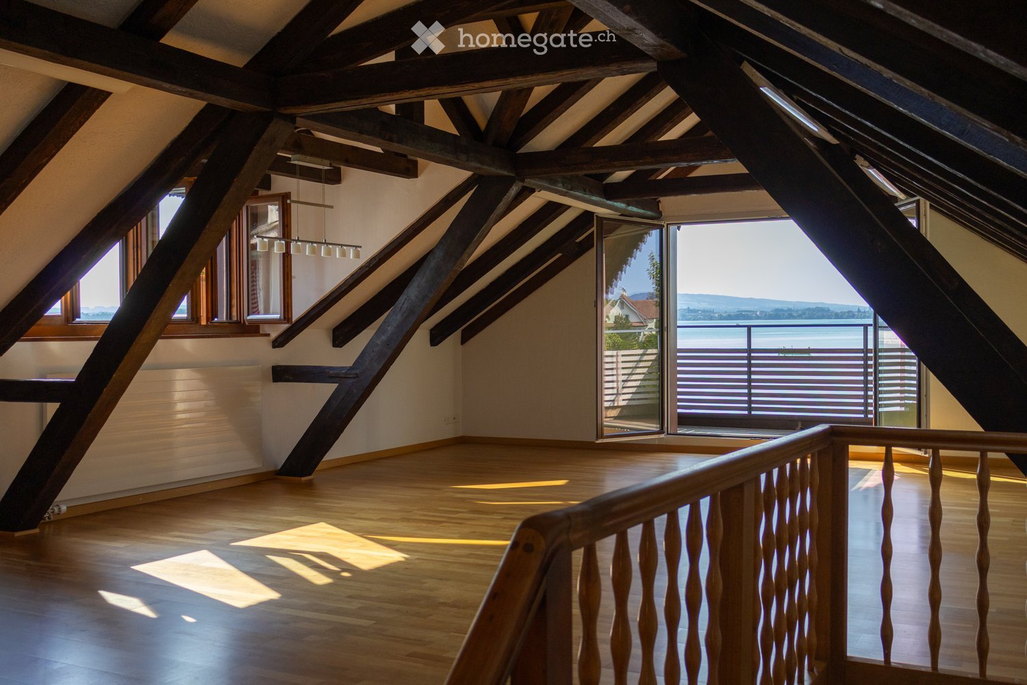 Bright, empty attic room with wooden beams, wooden floor, large windows, and a view of the lake and mountains.