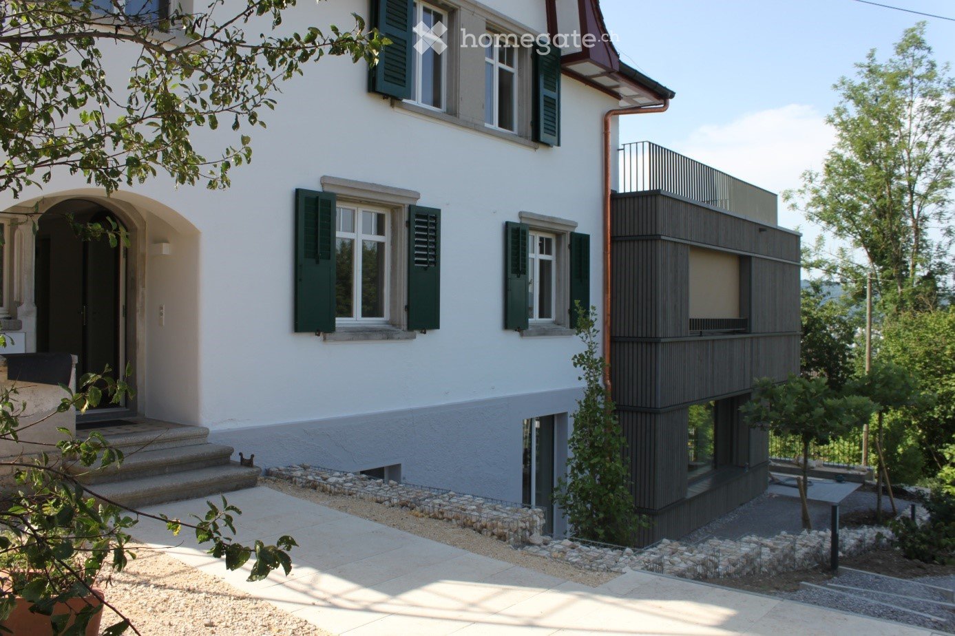 Two-story house with white exterior, green shutters, and a modern extension with a balcony. The entrance features steps leading to a door, and the surrounding area includes a paved walkway and small garden.