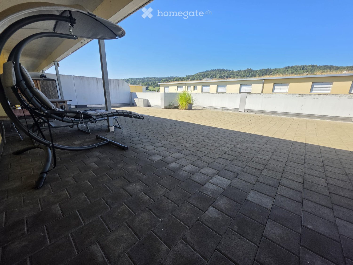 Roof terrace with dark tiled flooring, lounge chair, and a view of surrounding buildings and greenery.