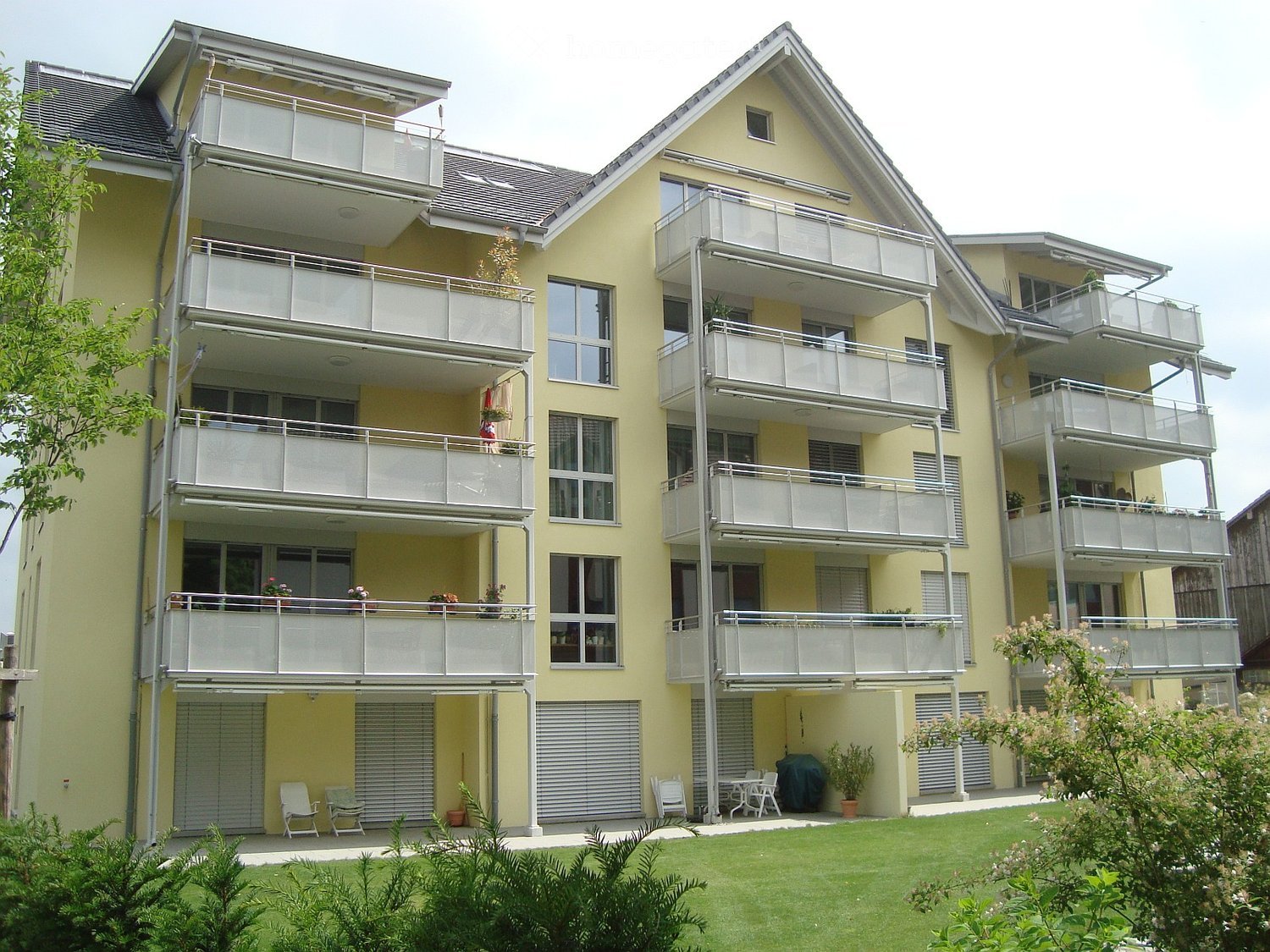 A modern yellow apartment building with multiple balconies, large windows, and a well-maintained garden area with chairs and tables.