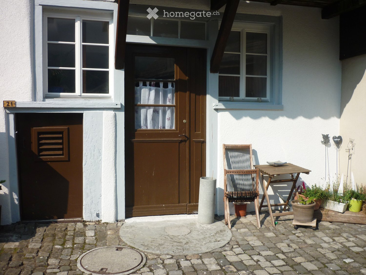 Entrance to a house with a small outdoor seating area featuring a wooden chair, table, and potted plants.