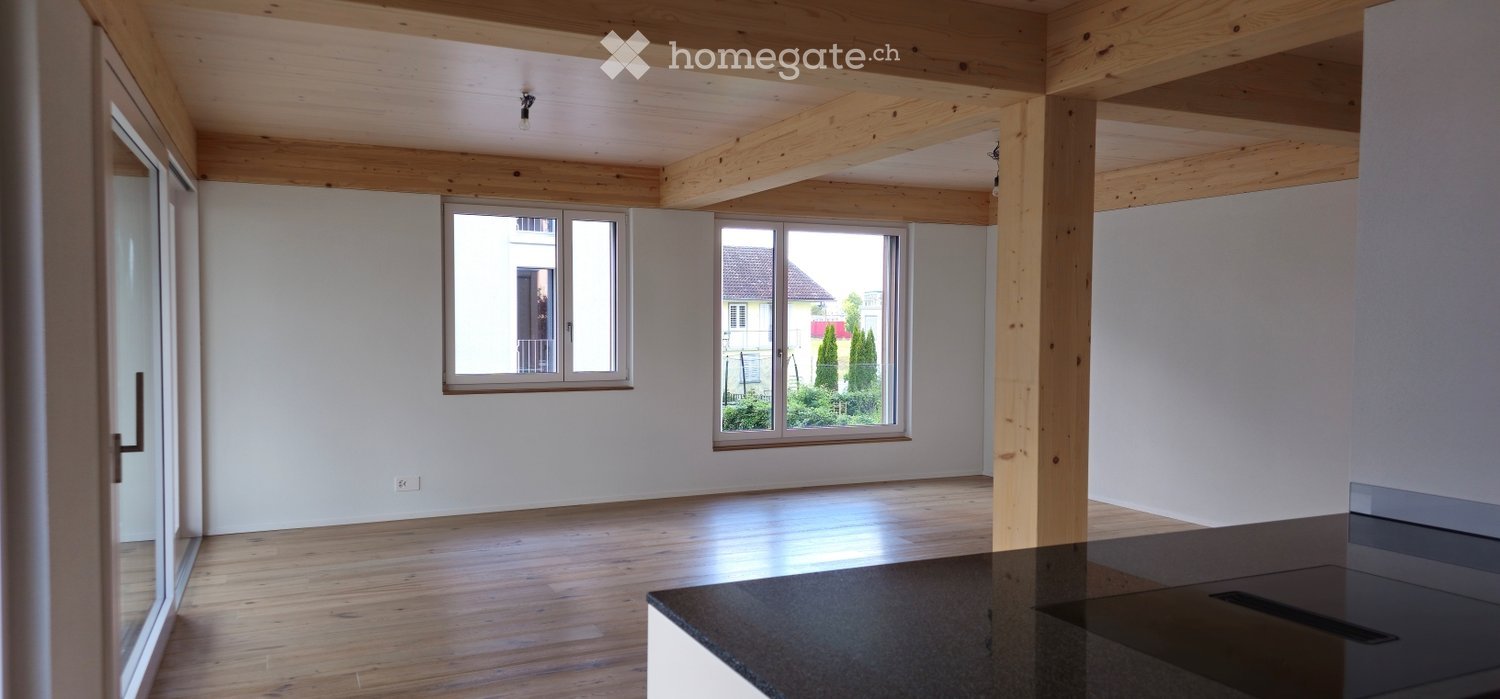 Bright, open room with wooden beams on the ceiling, large windows, and a polished wooden floor. The space is completely empty, with no furniture or decorations.