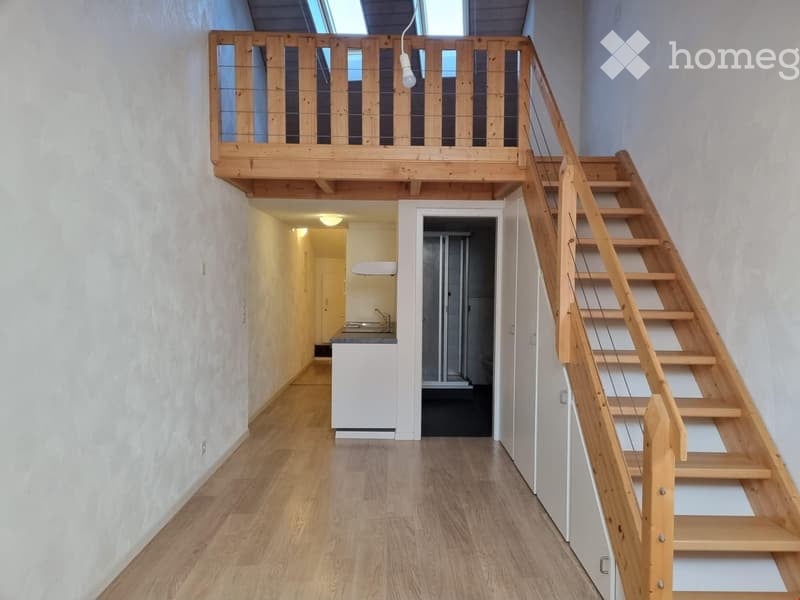 A narrow hallway with wooden flooring, leading to a small kitchen area and a staircase to an upper loft space. The loft has a wooden railing and skylight windows.