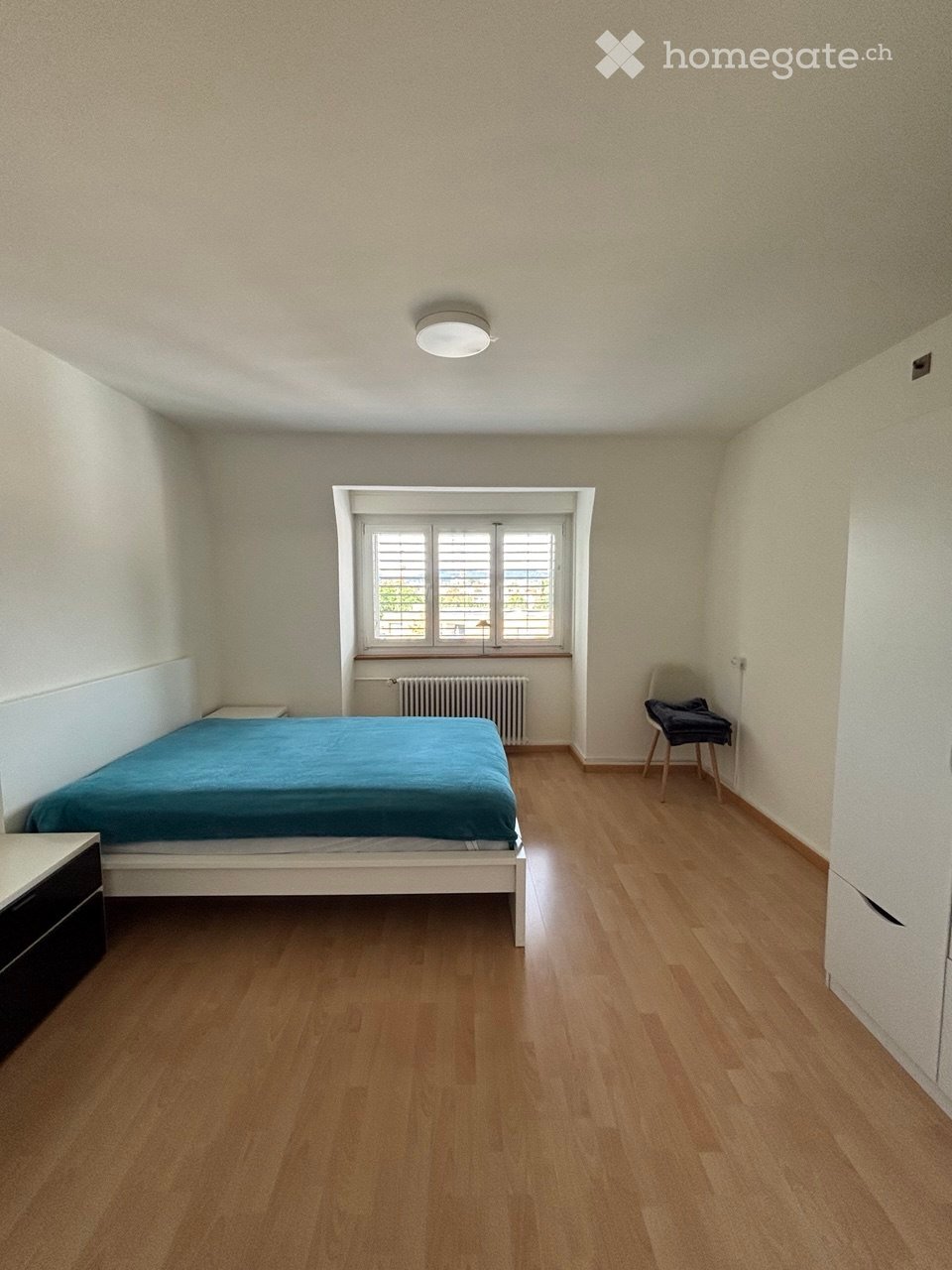 A furnished bedroom with a wooden floor, a single bed with blue bedding, a nightstand, a chair, and a radiator beneath a window.