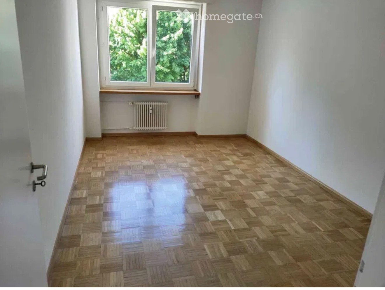 Empty room with wooden parquet flooring, white walls, and a window with a radiator below it.