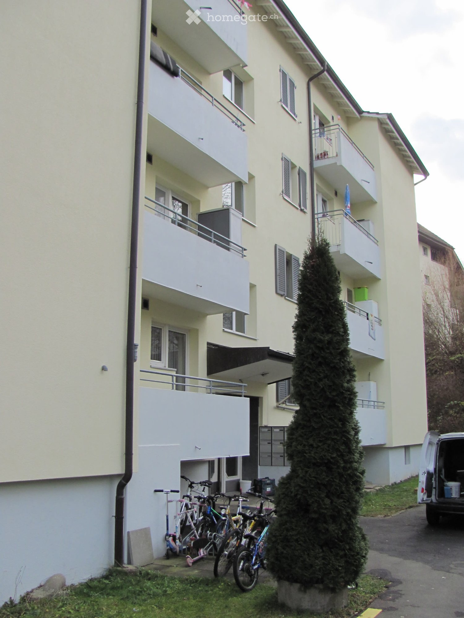 Multi-story apartment building with white exterior and balconies, bicycles parked in front, grass lawn