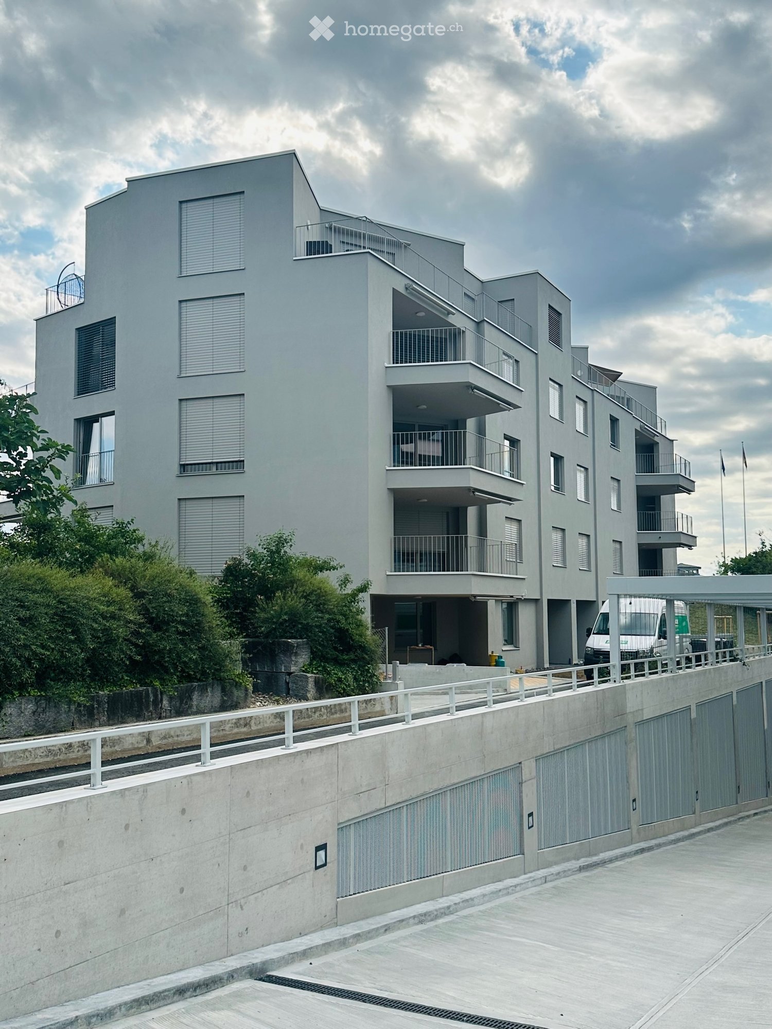 Modern multi-story residential building with grey facade, multiple balconies, and large windows. Features a raised ground floor with a concrete barrier and parking area.
