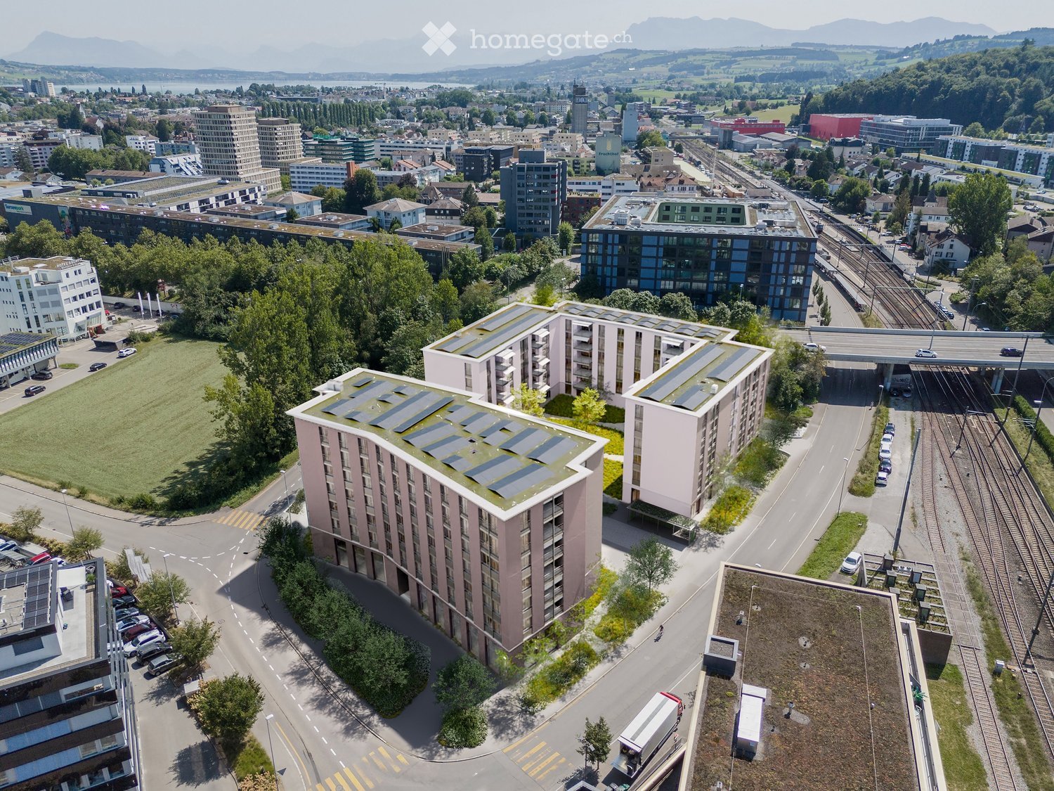 Aerial view of a modern residential complex with solar panels on the rooftops, surrounded by greenery, parking areas, and nearby urban infrastructure including roads, train tracks, and other buildings.