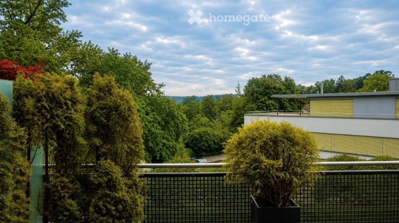Balcony with metal railing, potted plants, and a view of a modern building and surrounding greenery.