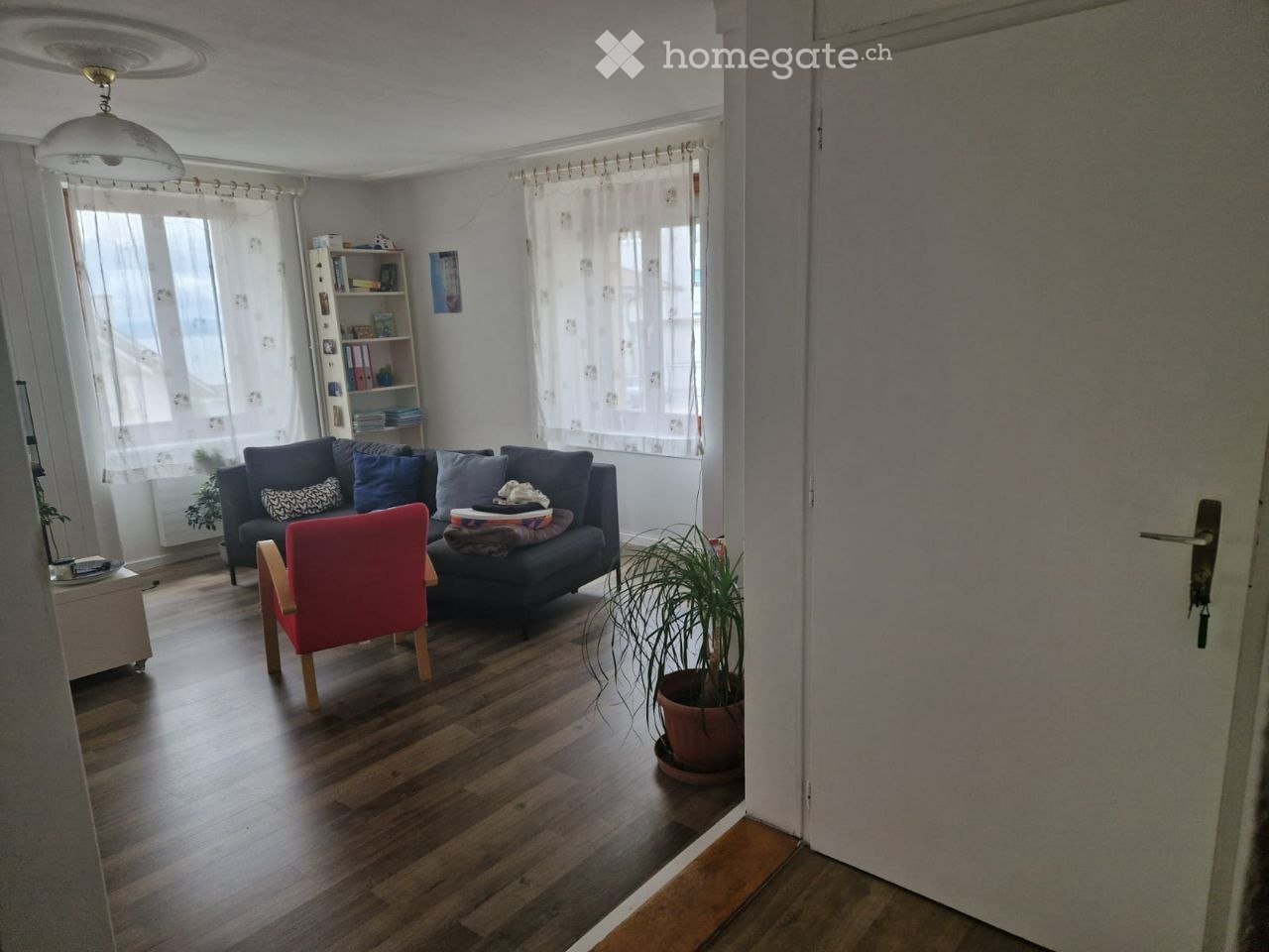 Living room with wooden flooring, large windows with white curtains, a dark blue sofa with pillows, a red wooden chair, a potted plant, and a bookshelf.