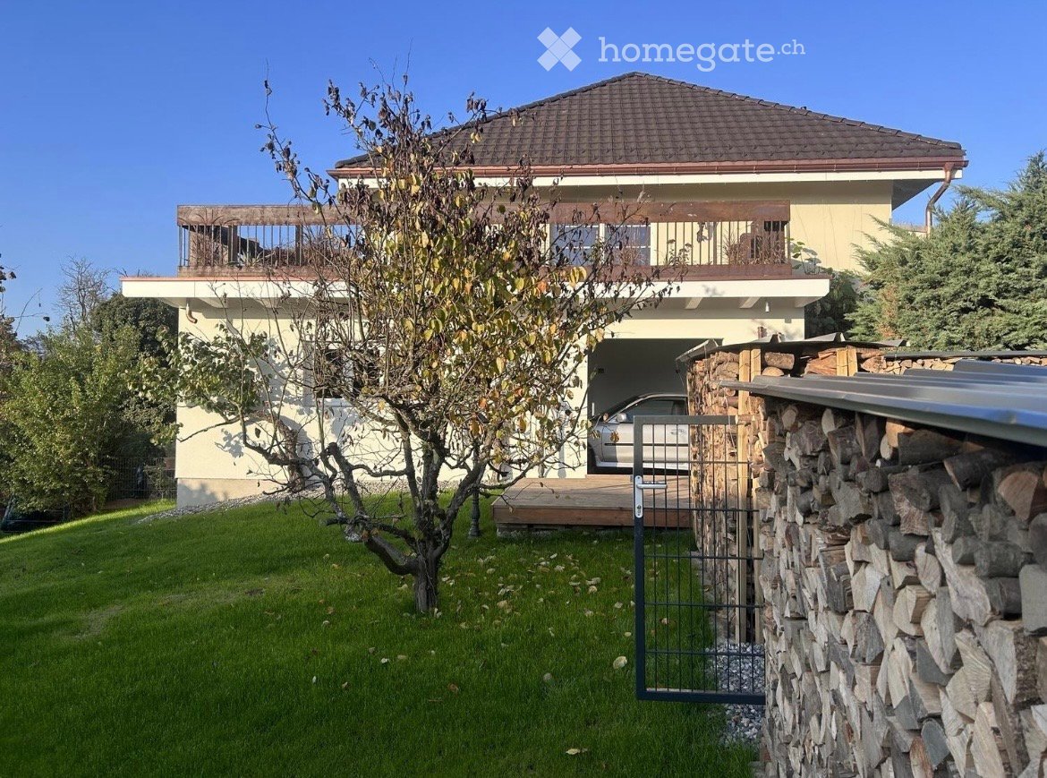 Two-story house with a garage, balcony, and a stone wall entrance. The house has a beige exterior with a tiled roof and is surrounded by a well-maintained lawn and trees.