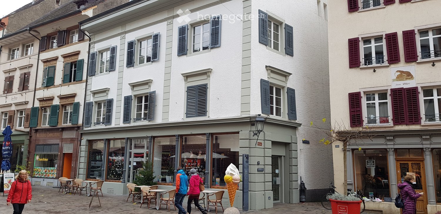 A row of multi-story buildings with classic European architecture, featuring shutters, large windows, and storefronts. The ground floor includes cafes and shops with outdoor seating areas.