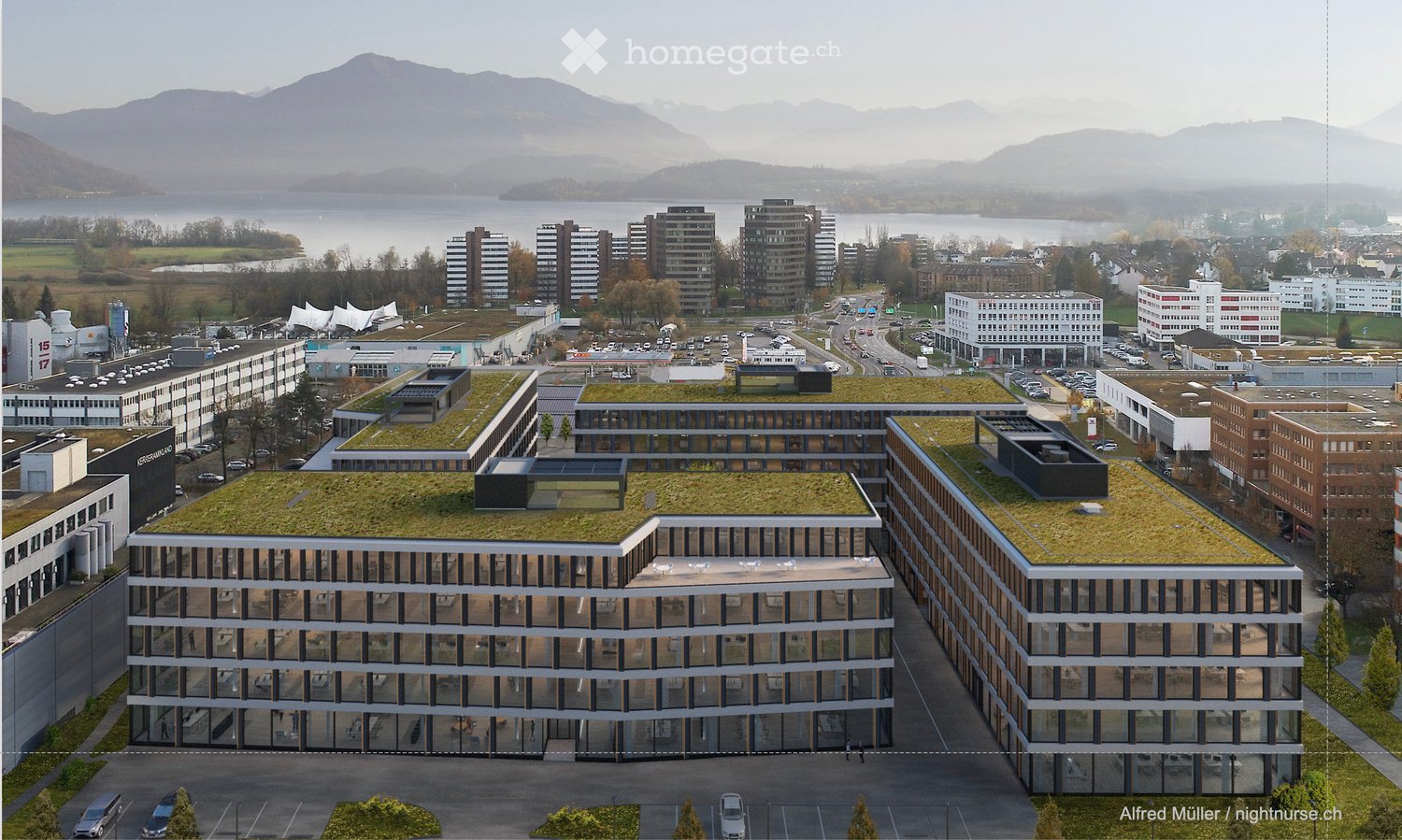 A modern multi-story building with a green roof, surrounded by other structures and a parking lot, with a scenic lake and mountains in the background.