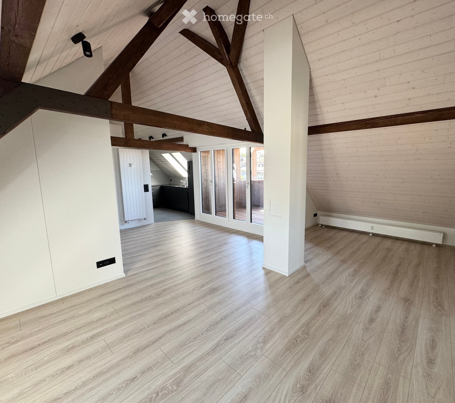 Bright, open attic space with wooden beams, light wooden flooring, and large glass doors leading to a balcony. The room is completely empty with no furniture or equipment.