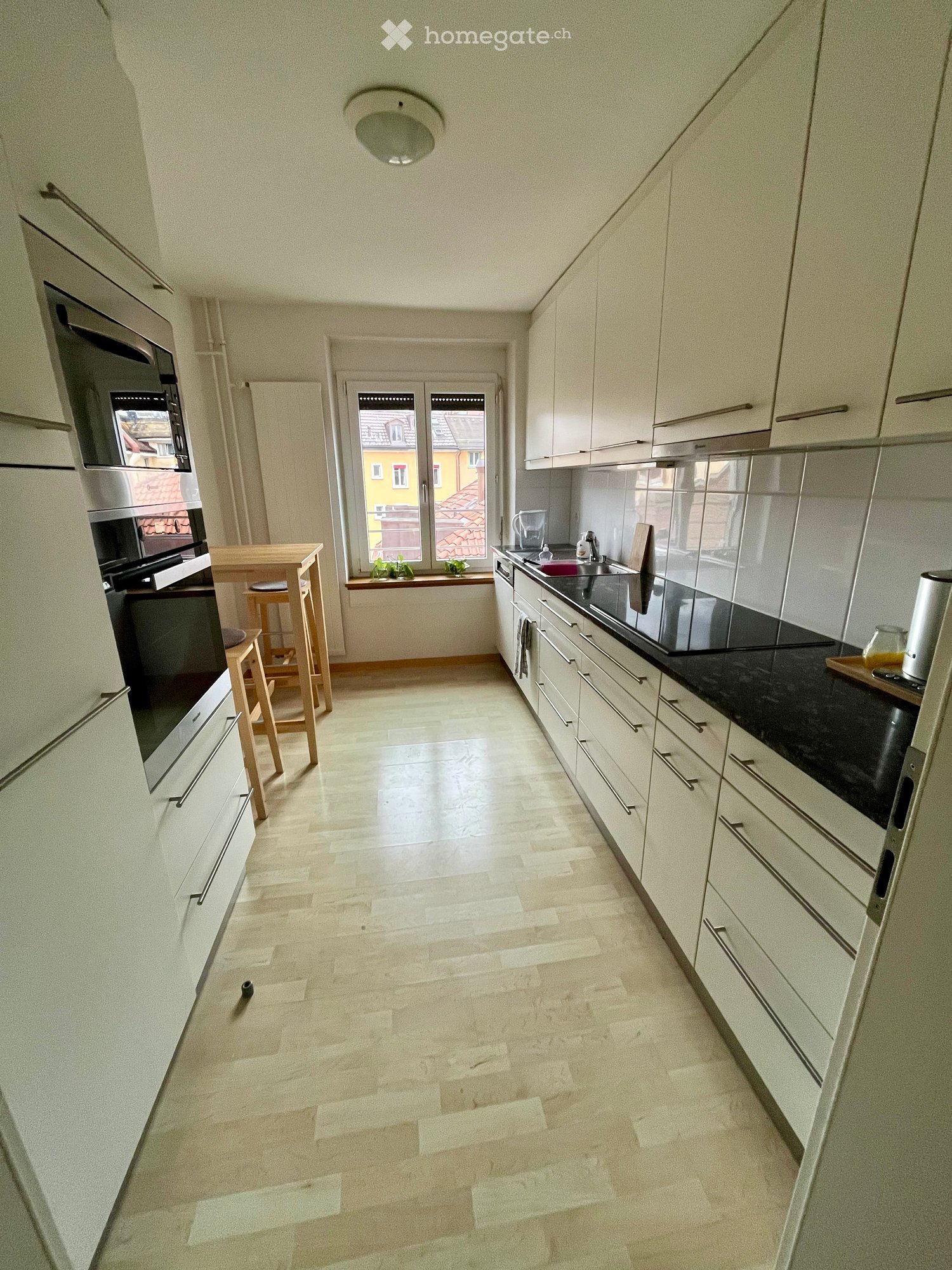 Modern kitchen with white cabinets, black countertops, and a central island. Features include a sink, oven, microwave, and a small dining area with stools.