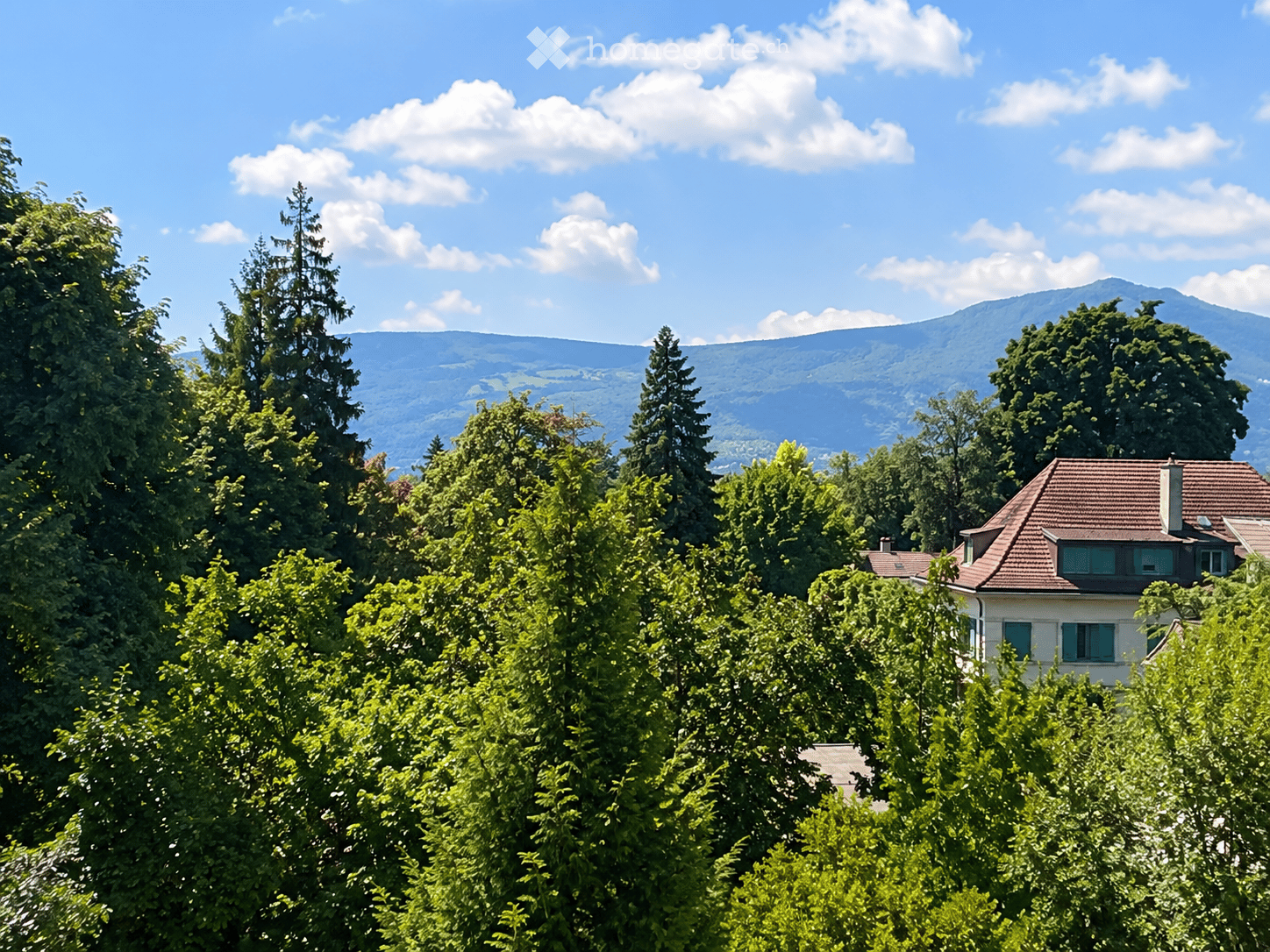 A two-story house with a red-tiled roof and green shutters, surrounded by lush green trees and a scenic mountain backdrop.