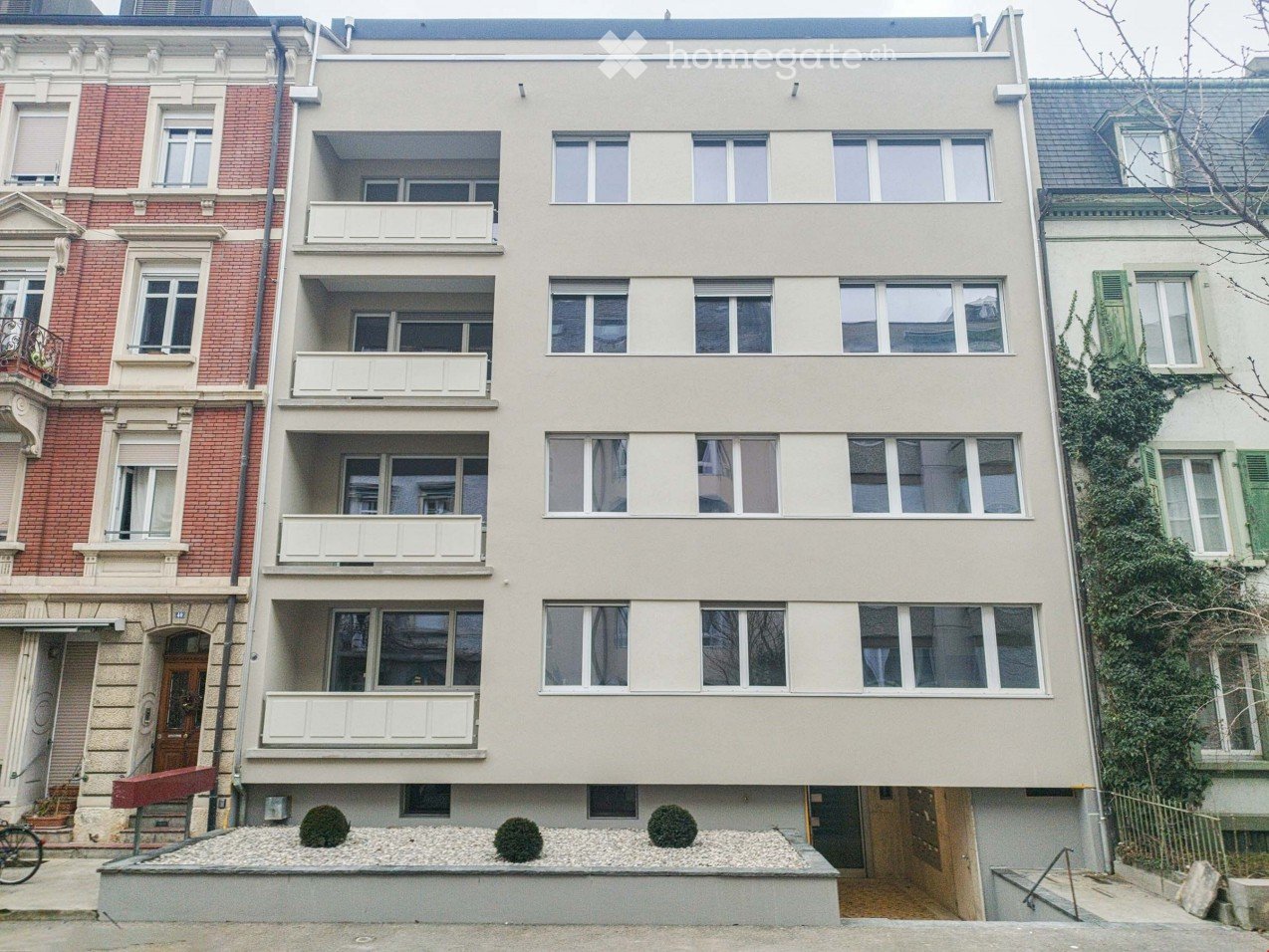 Four-story residential building with beige facade, multiple windows, and white balconies. Adjacent buildings include red brick and green ivy-covered structures. Small landscaped area with gravel and shrubs in front.