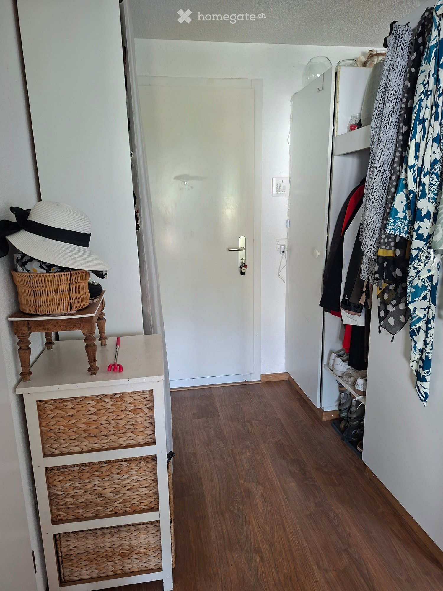 Hallway with wooden flooring, white walls, a small wooden table with a woven basket and hat, a coat rack with clothes, and a door leading to another room.