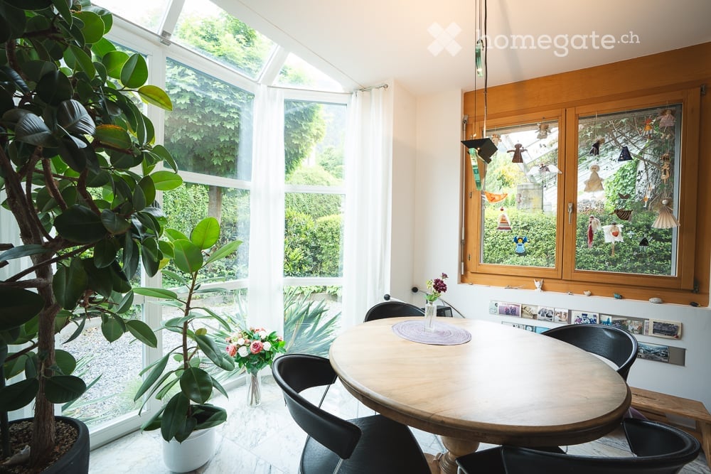 Bright, airy room with large windows and a skylight, featuring a round wooden dining table surrounded by black chairs, potted plants, and a vase with flowers on the table.