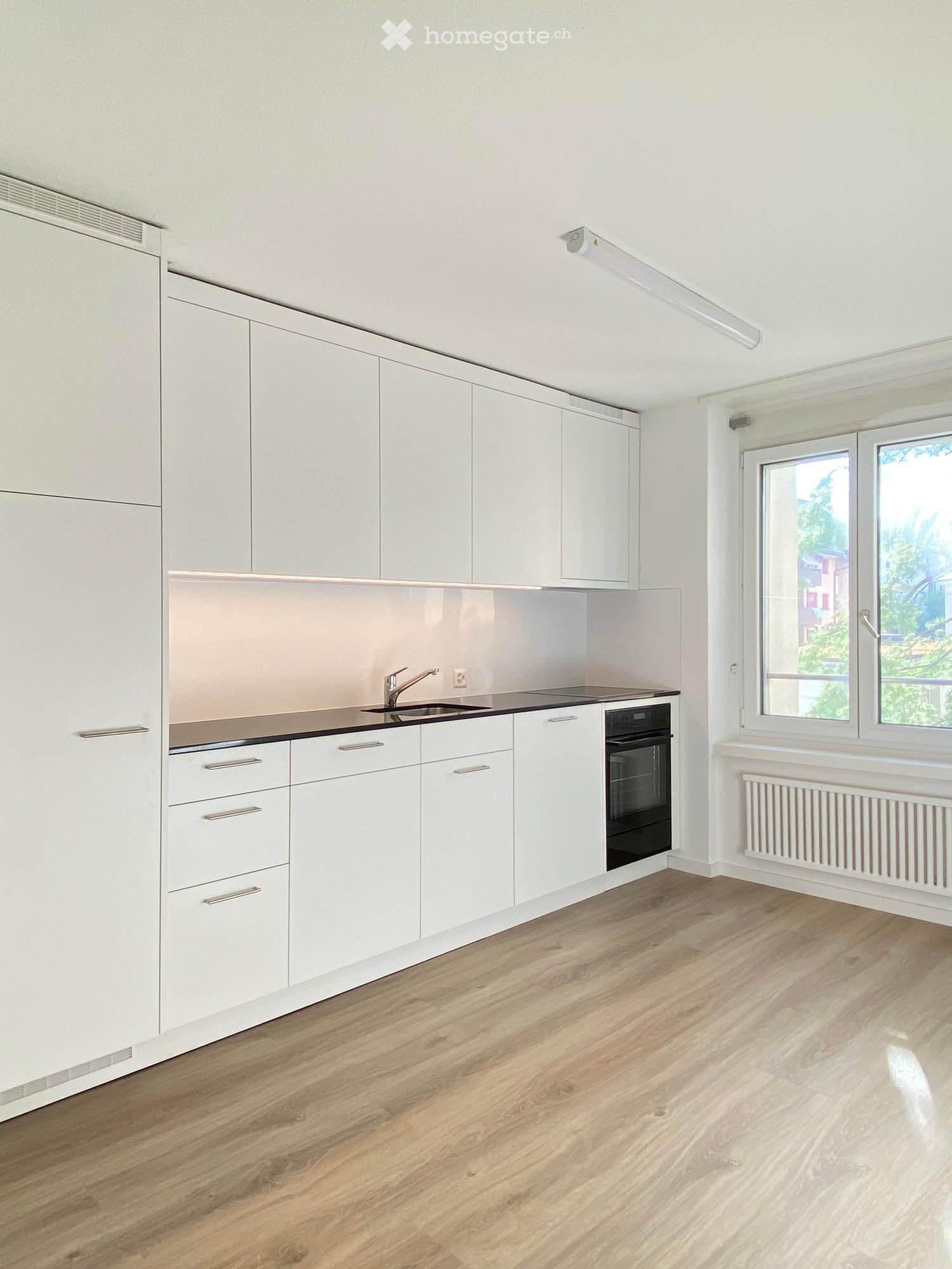 Modern kitchen with white cabinets, wooden flooring, and a window overlooking greenery.