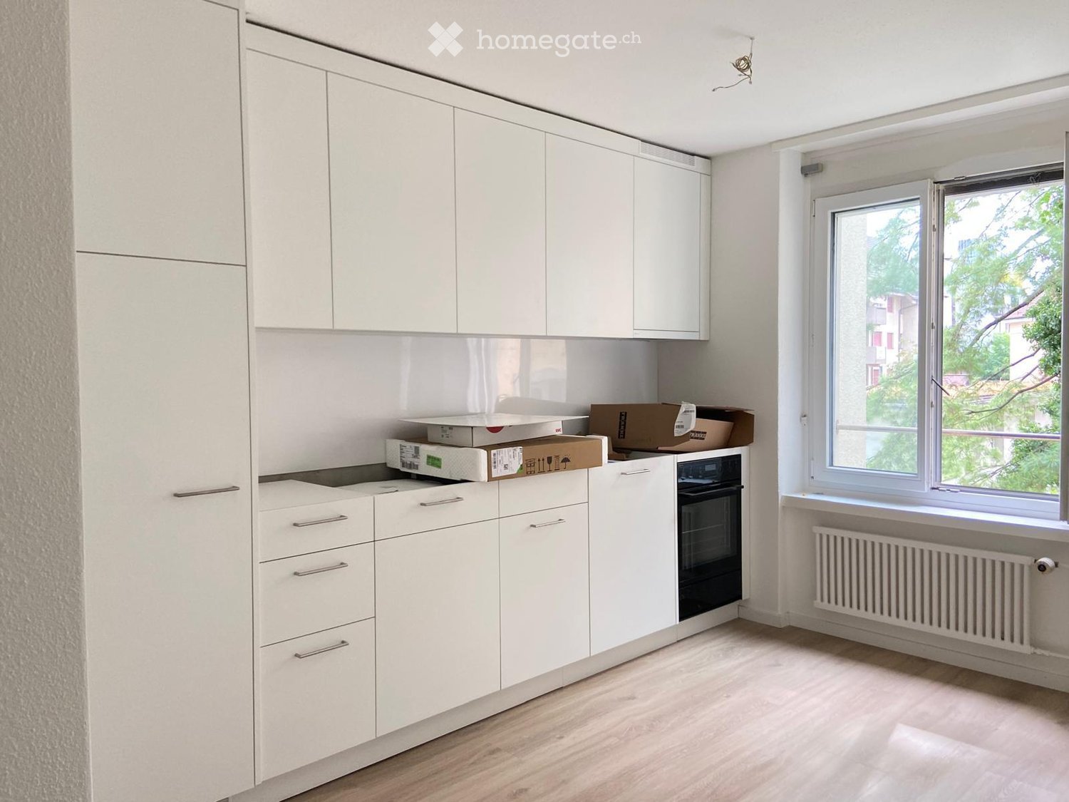 Modern white kitchen with fitted cabinets, an oven, and a window overlooking greenery.