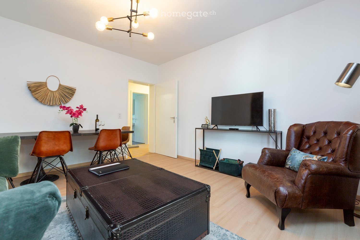 Modern living room with a leather armchair, coffee table, TV, and bar area with stools. Features a chandelier, decorative wall art, and a small plant on the bar.