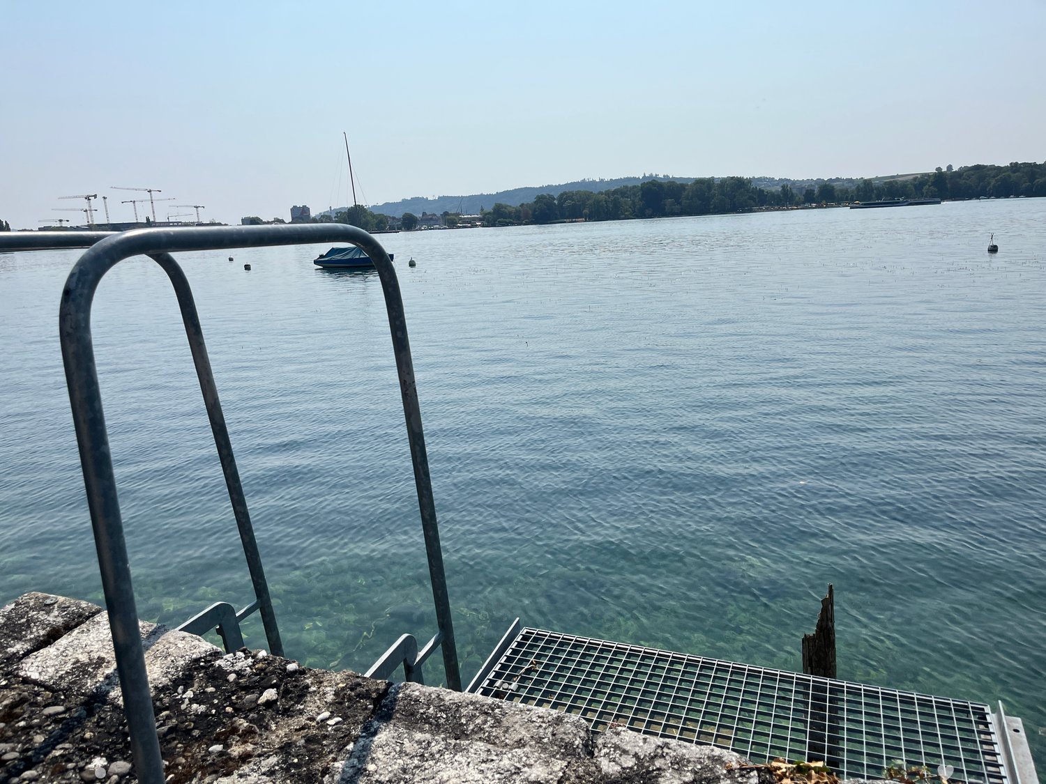 View of a calm lake with a small boat, distant construction cranes, and a distant shoreline with trees and buildings.