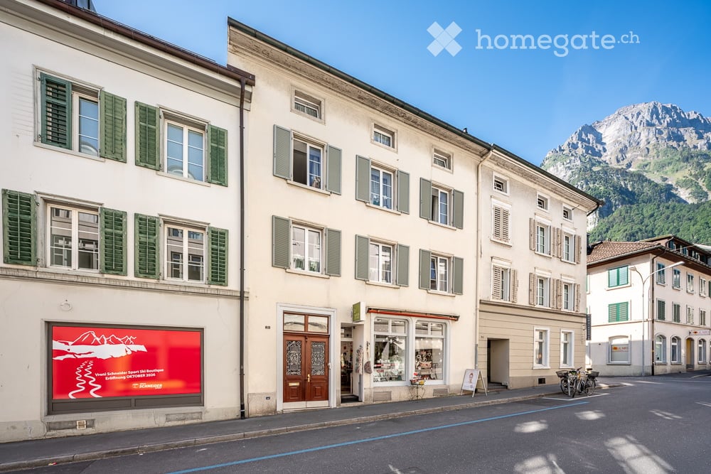 A row of traditional European buildings with white facades, green shutters, and large windows, featuring a storefront with a red signboard and bicycles parked outside, with a mountain view in the background.