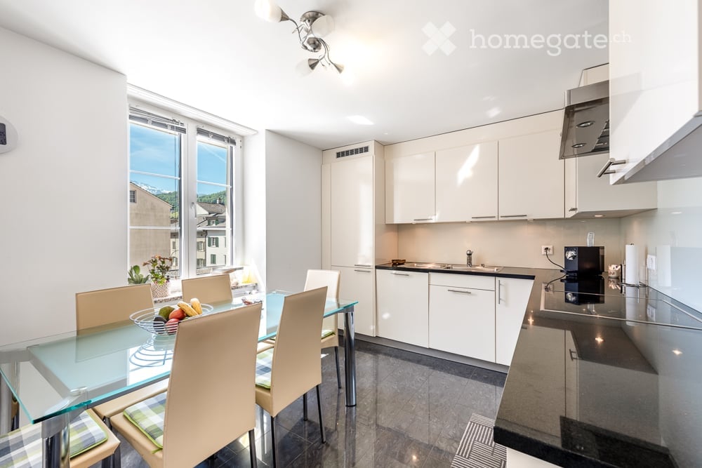 Modern open-plan kitchen and dining area with white cabinets, stainless steel appliances, a glass dining table with four chairs, and a fruit bowl on the table. Large windows offer a scenic view.