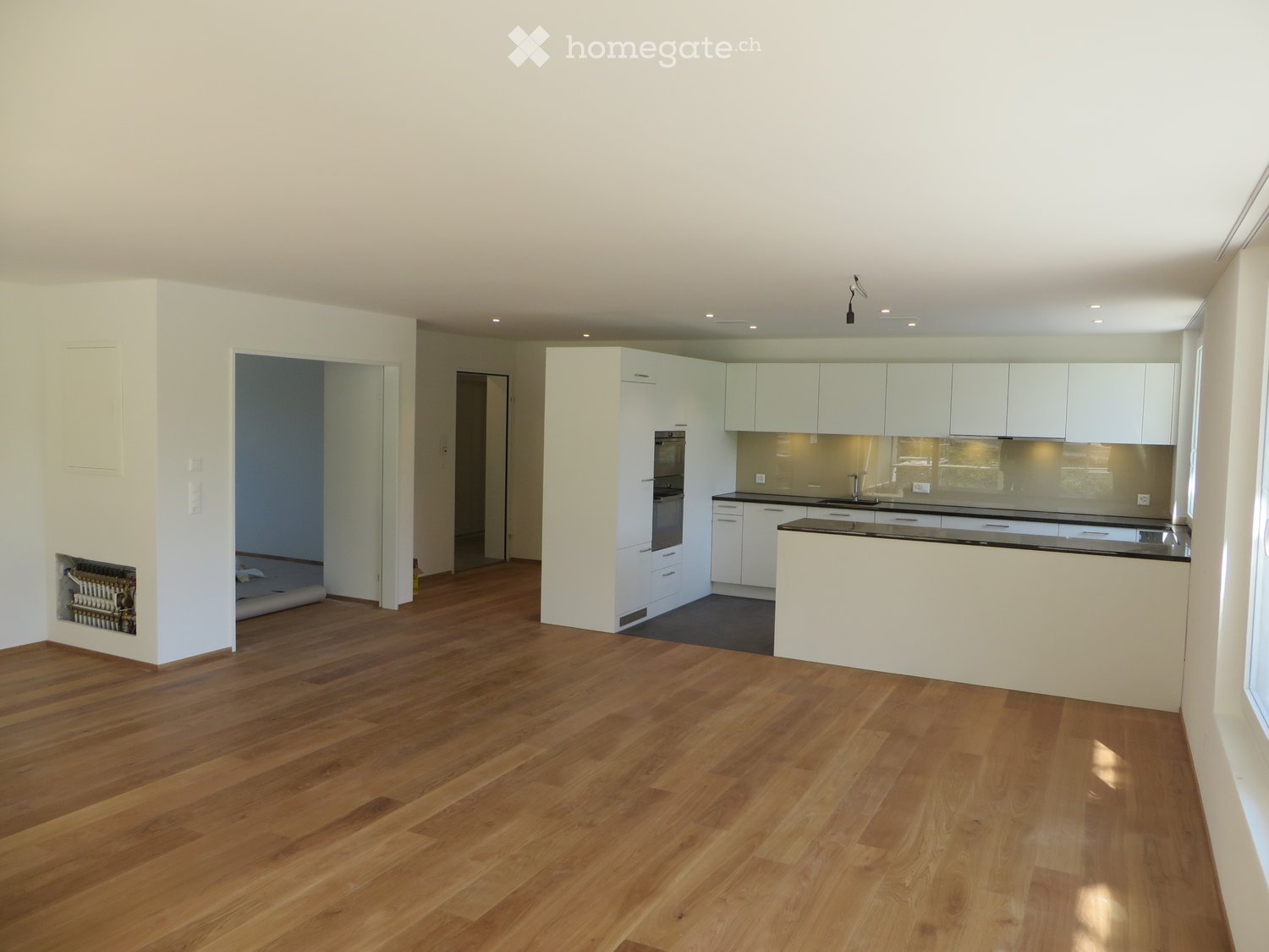 Open-plan space with light wooden flooring, white cabinetry, and a modern kitchen area featuring built-in appliances and a central island.
