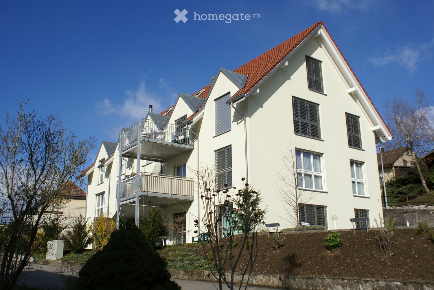 Modern two-story house with white exterior, red-tiled roof, multiple windows, and a balcony on the upper level.