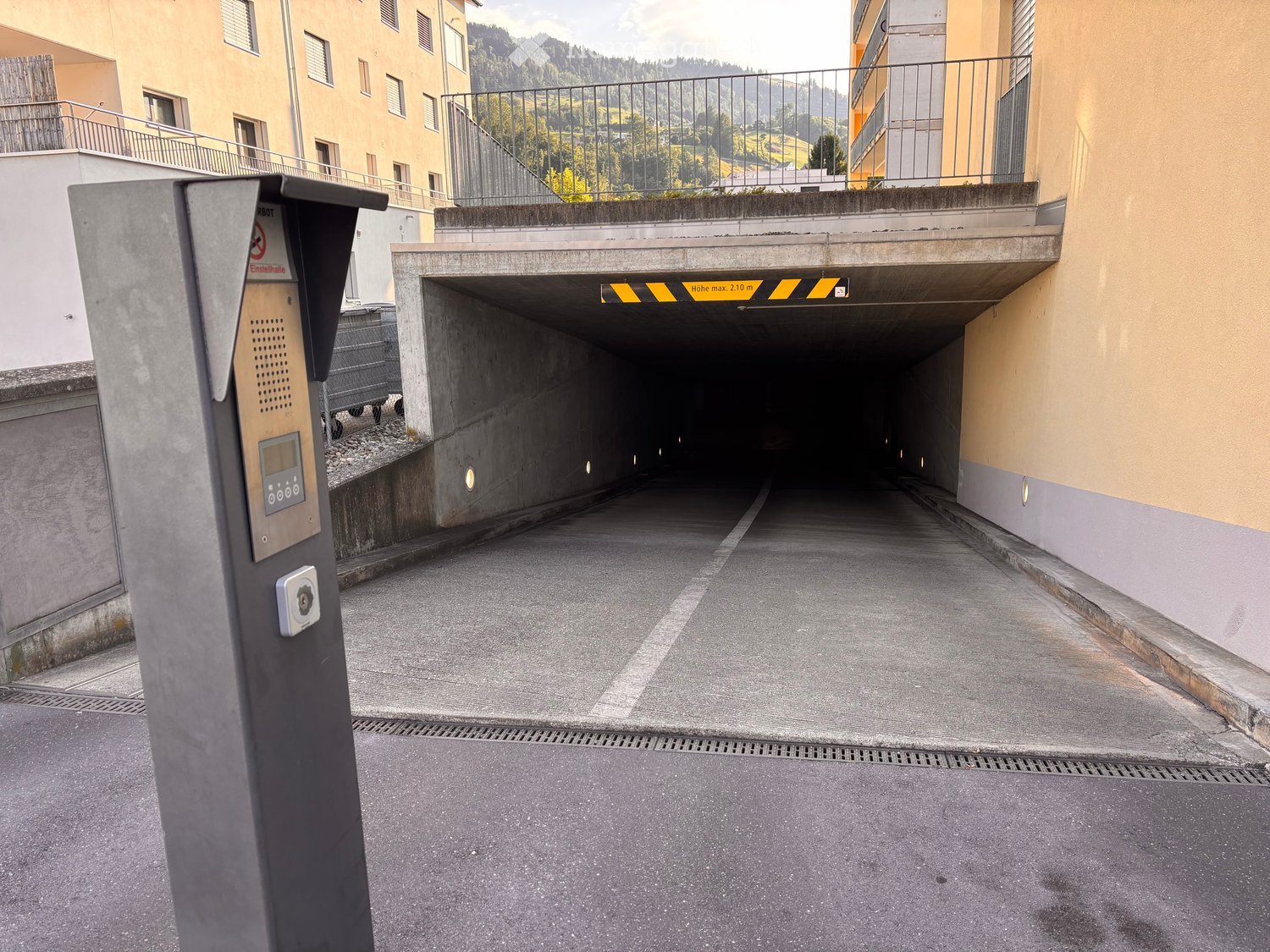 Entrance to a multi-story building with a covered parking area, featuring a concrete ramp and a security access point.
