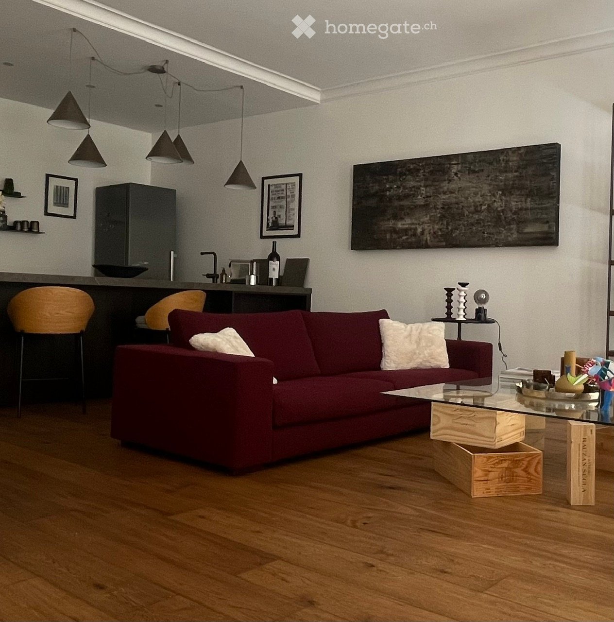 Modern living room with a red sofa, glass coffee table, pendant lights, and a small bar area with stools and a cabinet.