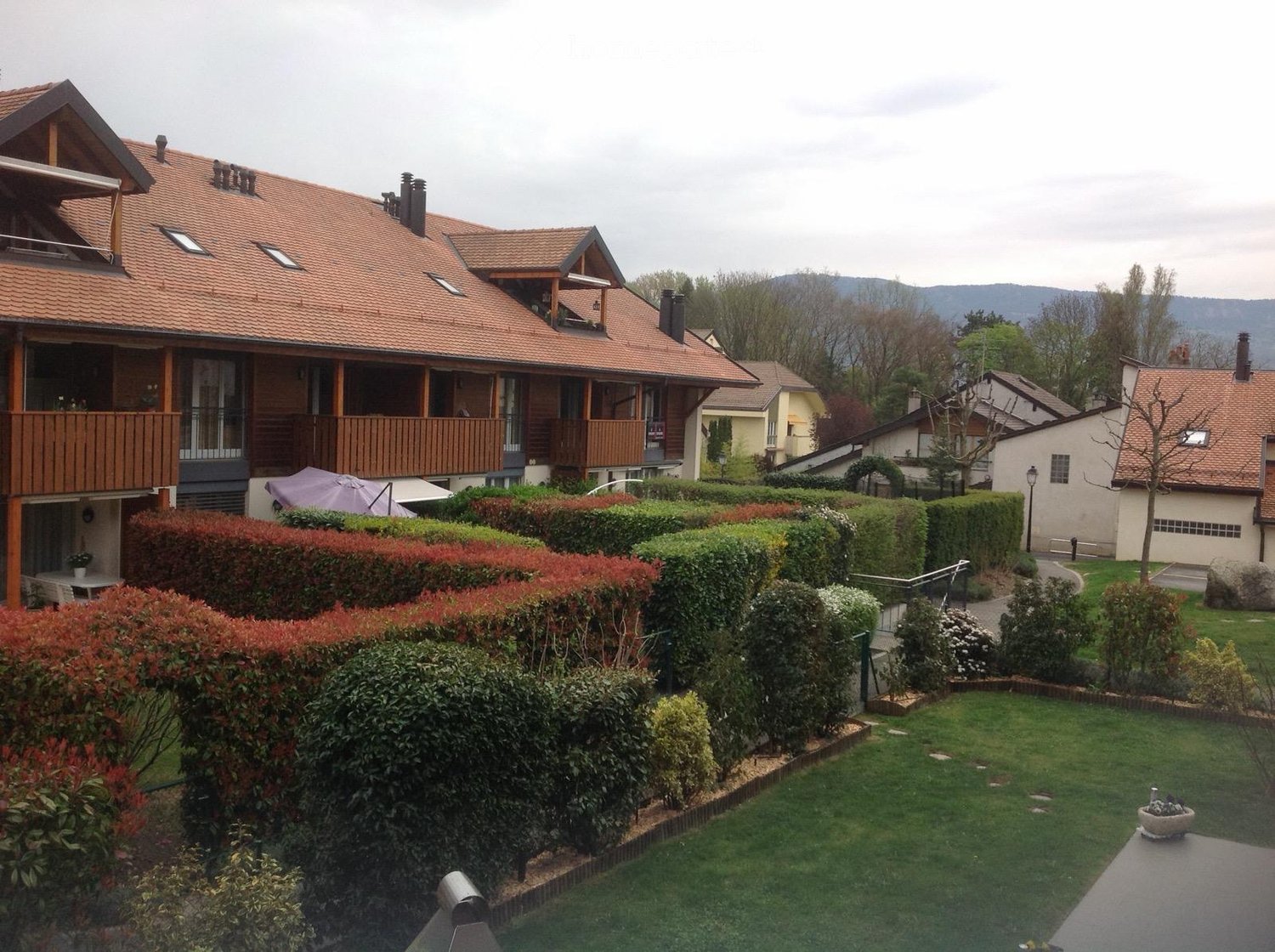 Well-maintained garden with neatly trimmed hedges, a small patio area, and a wooden deck on the house. The house features a red-tiled roof, large windows, and a balcony.
