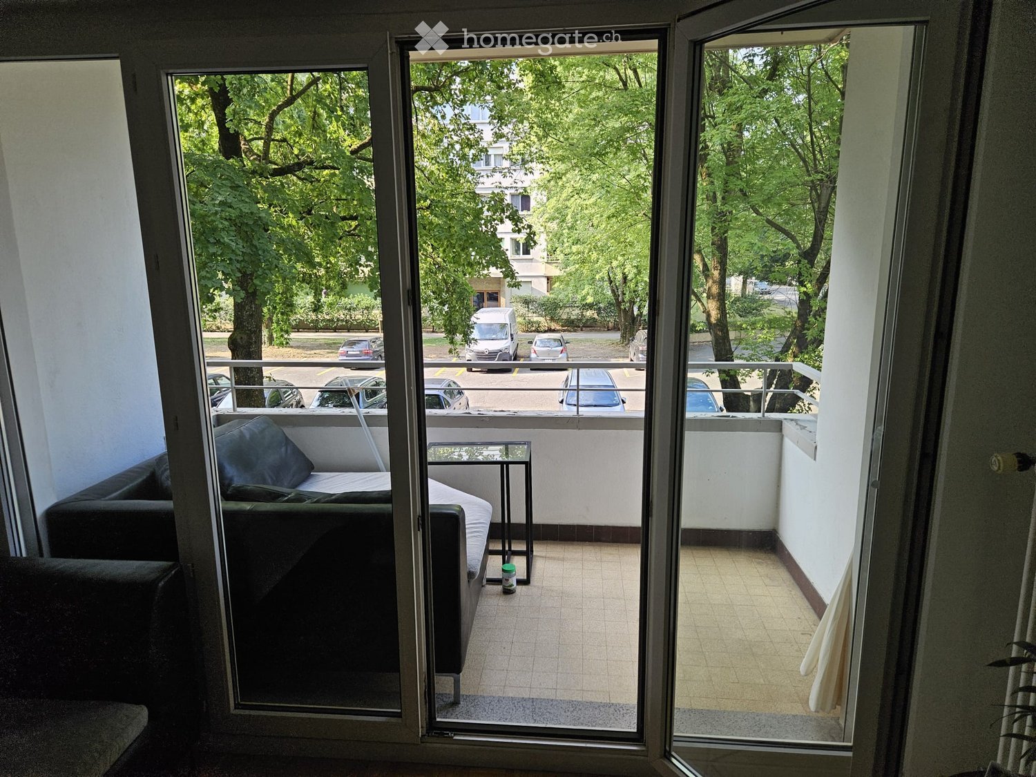 Balcony with tiled floor, white walls, metal railing, sofa, small table, and a view of a parking lot with cars and trees.