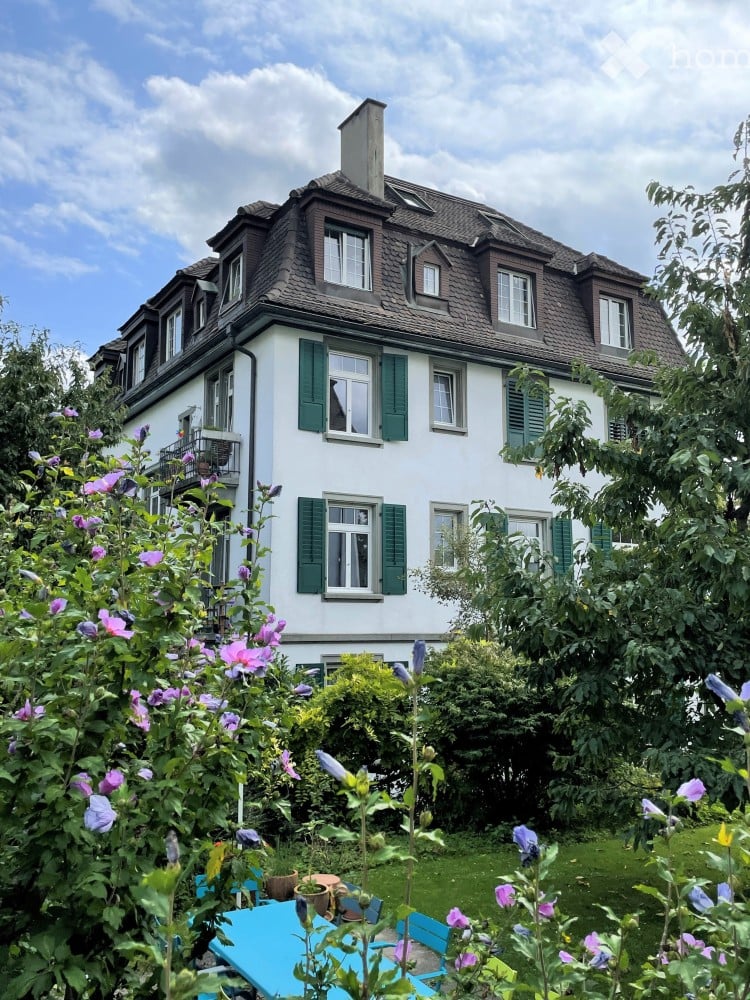A two-story white house with green shutters and a pitched roof, surrounded by a garden with colorful flowers and outdoor furniture.