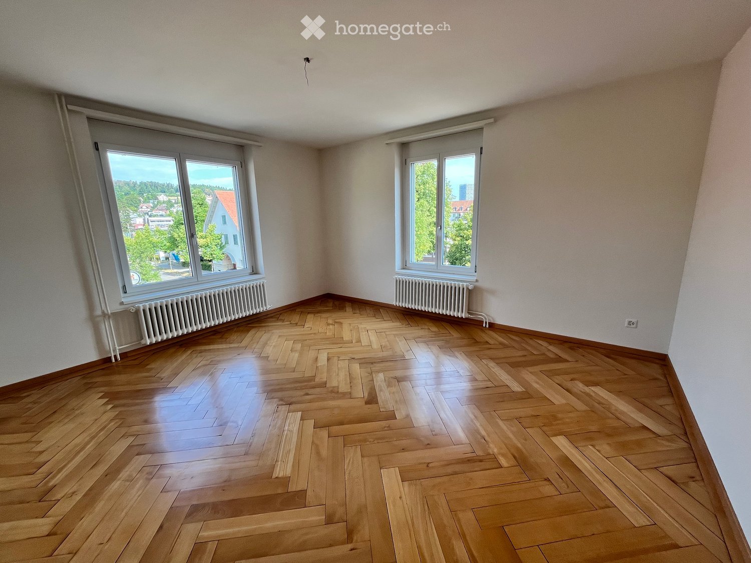 Empty room with wooden herringbone flooring, white walls, large windows with blinds, and radiators beneath the windows.