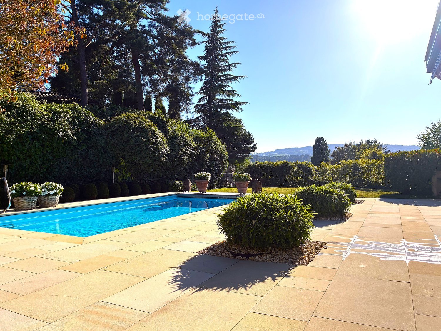 A well-maintained garden area with a swimming pool, surrounded by lush greenery, trees, and neatly arranged potted plants. The pool is surrounded by a tiled patio area, and the background features a scenic view of hills and trees.