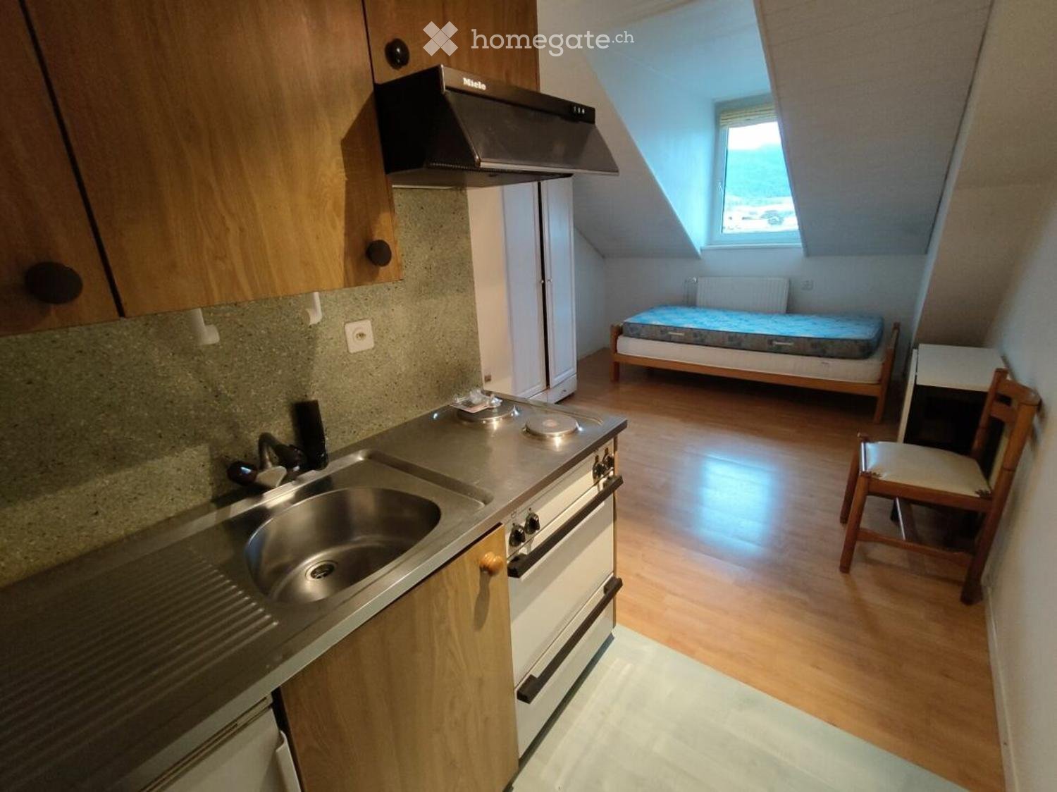 Compact kitchen area with sink, stove, and refrigerator, connected to a small bedroom with a bed and chair.