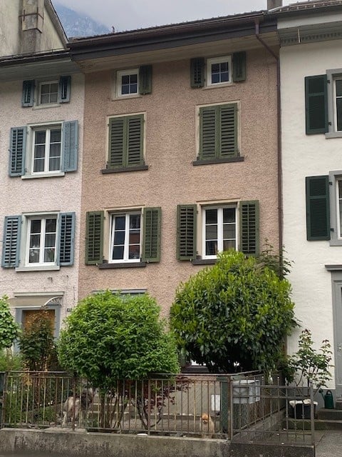 A three-story residential building with light pink facade, green shutters, and multiple windows. Small trees and a metal fence are visible in the foreground.