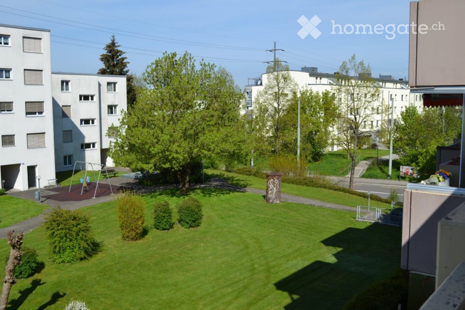 A well-maintained garden area with green grass, small shrubs, and trees, surrounded by residential buildings and a playground.